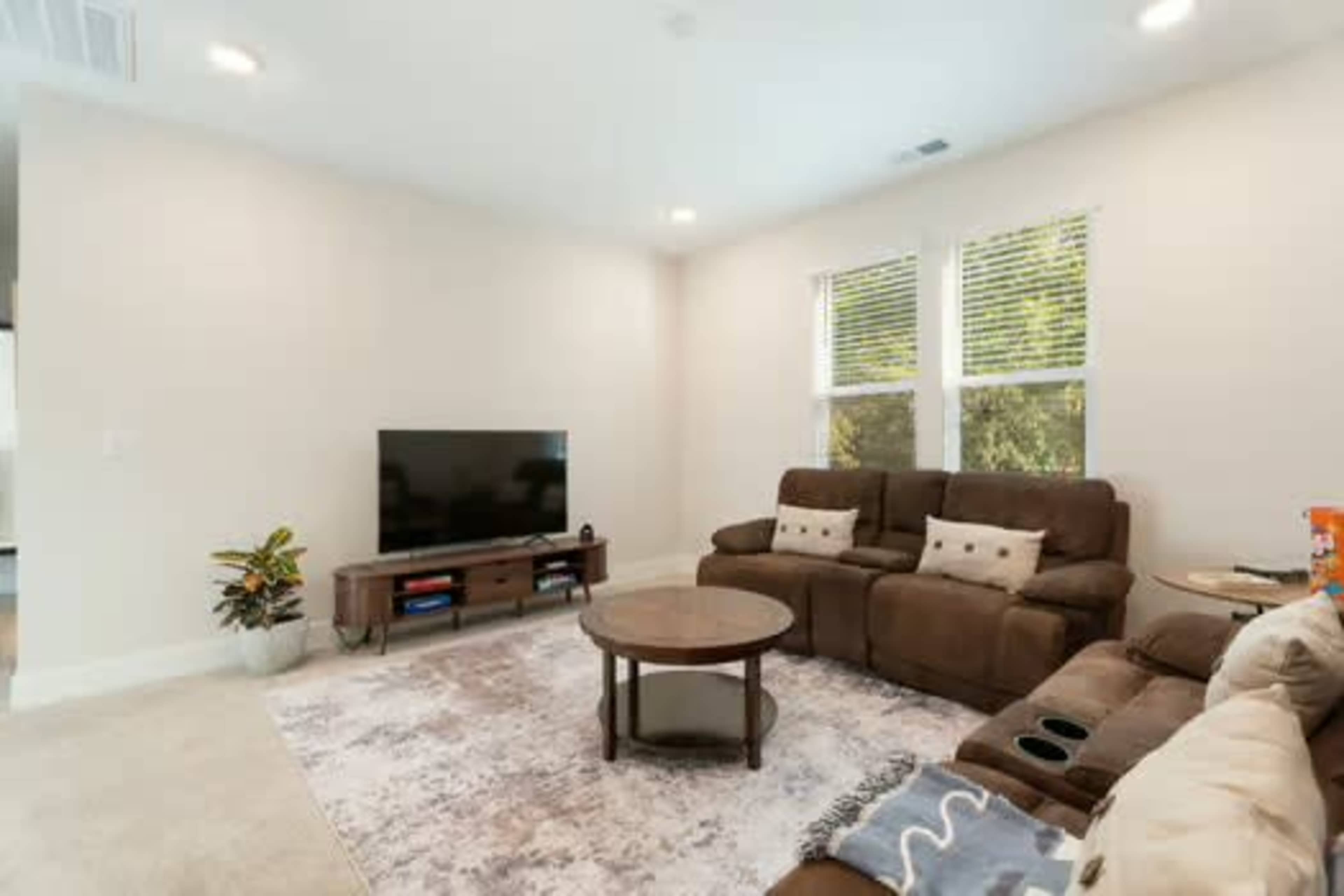 The living room features a brown sectional sofa, a round coffee table, a television on a wooden stand, and windows allowing natural light.
