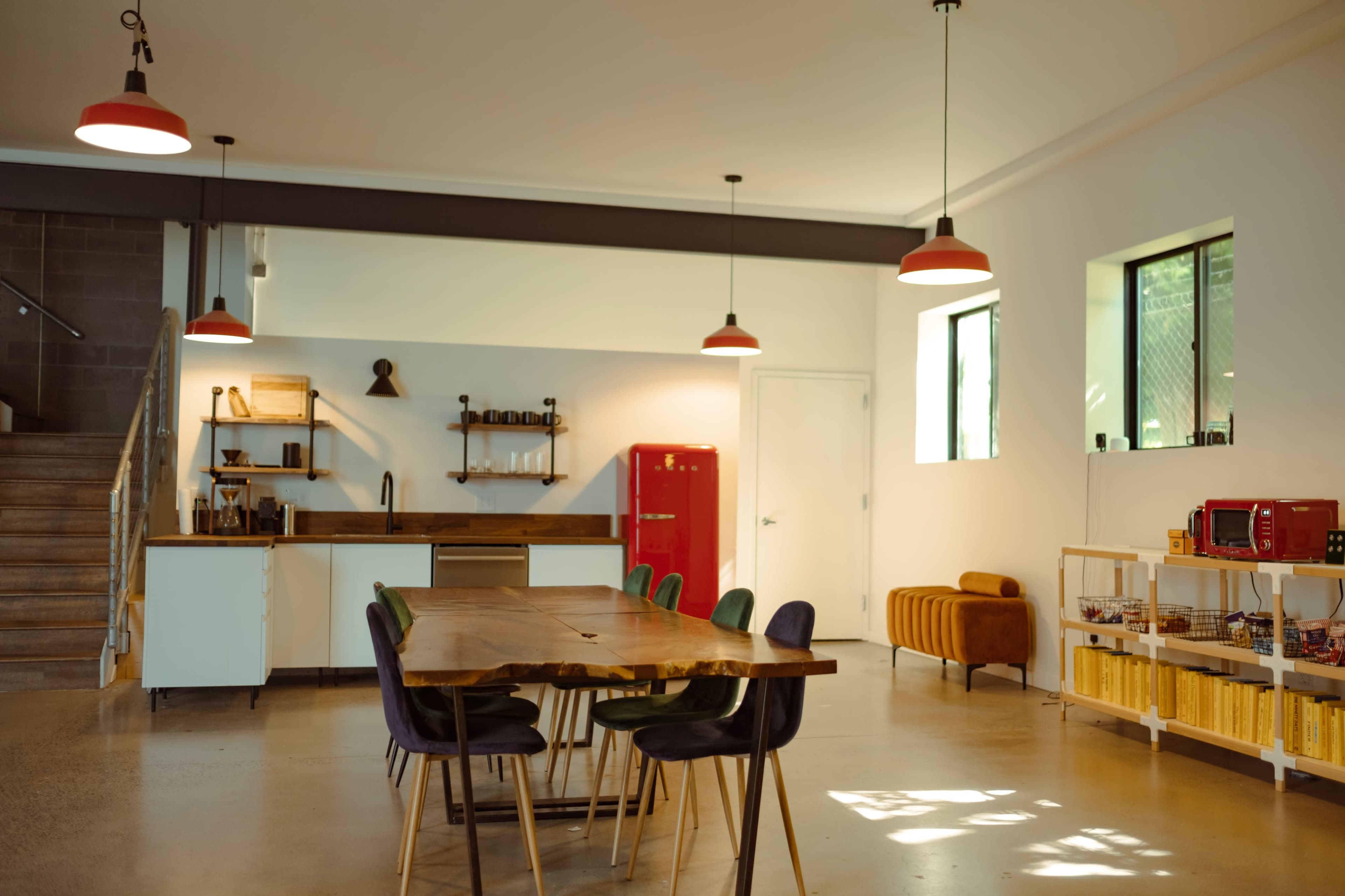 A modern kitchen and dining area features a large wooden table surrounded by colorful chairs, a red refrigerator, and various appliances on a countertop.