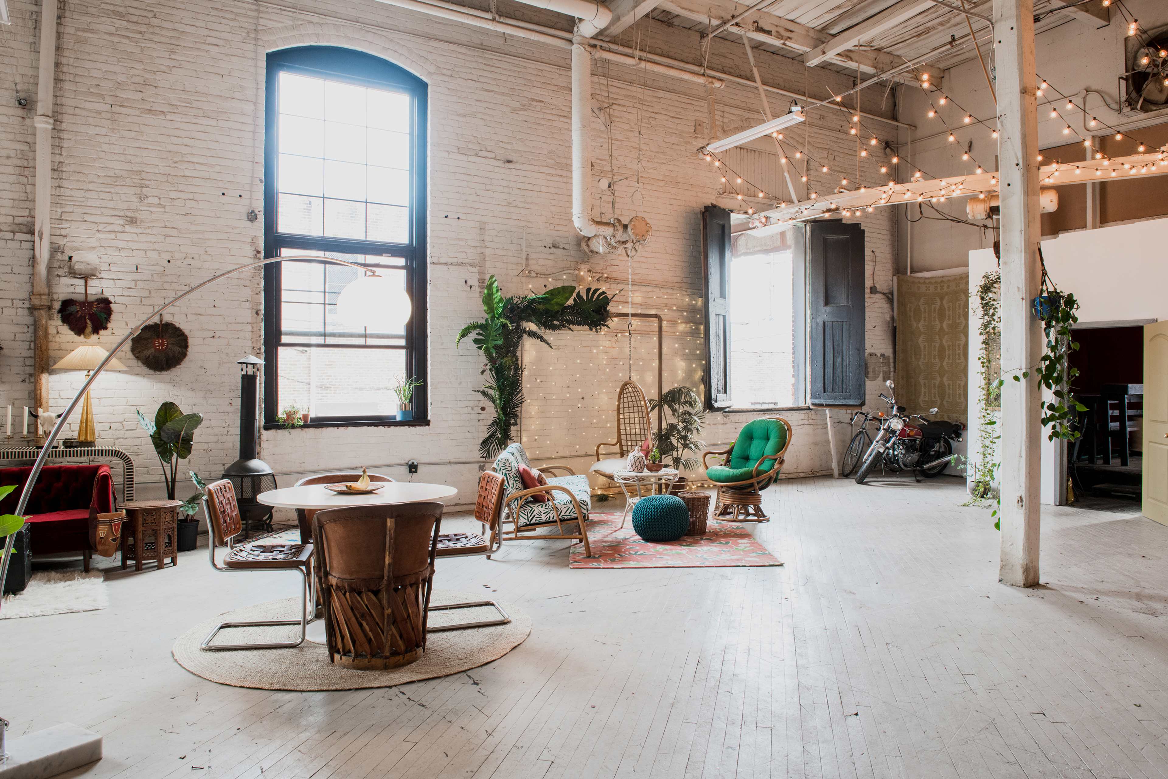 The spacious room features eclectic furniture, including a round table and various seating arrangements, with string lights and plants creating a vibrant atmosphere.