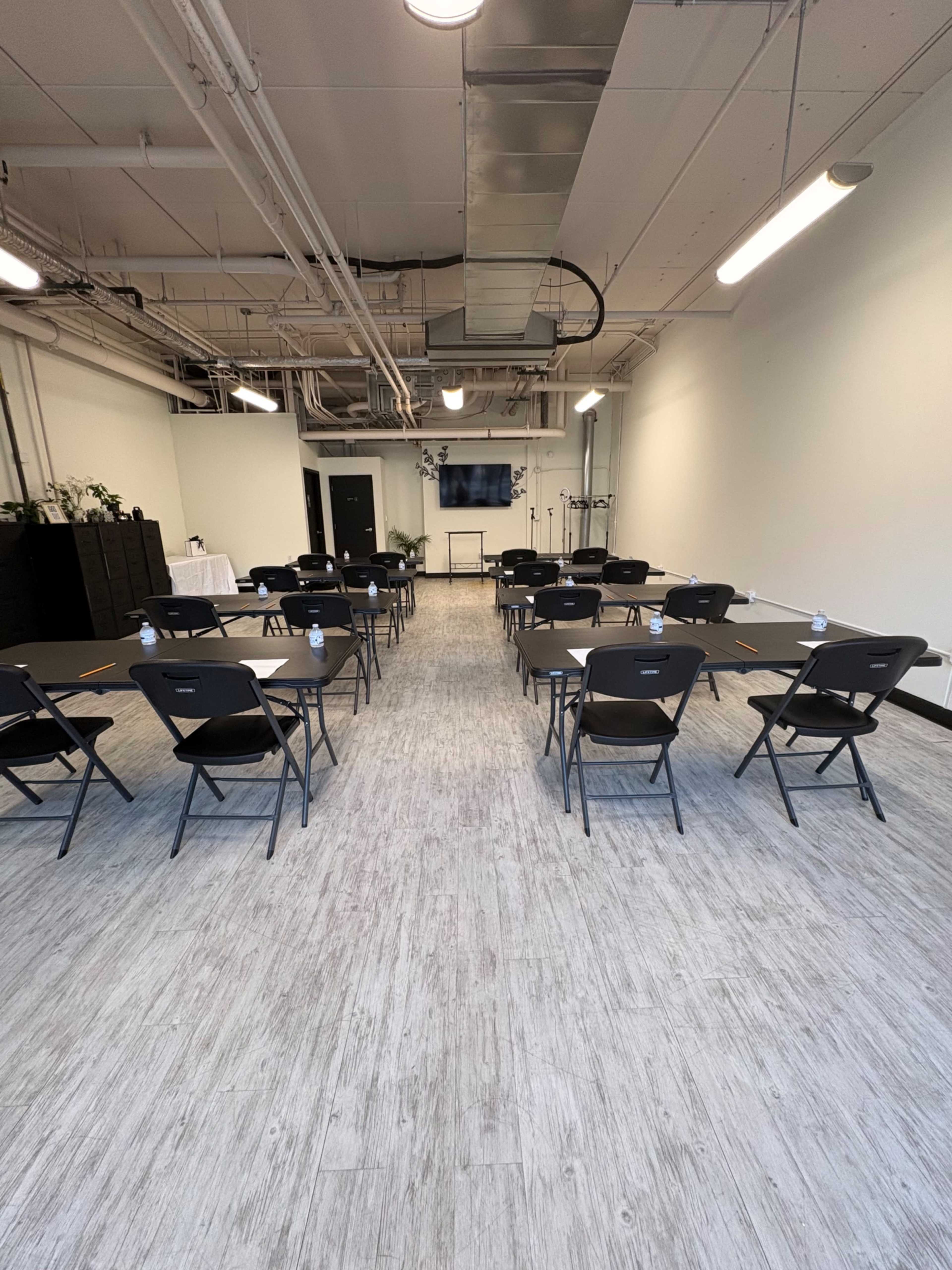 The image shows a spacious room set up for a meeting, featuring rows of folding chairs facing a presentation screen, with light-colored flooring and minimal decor.