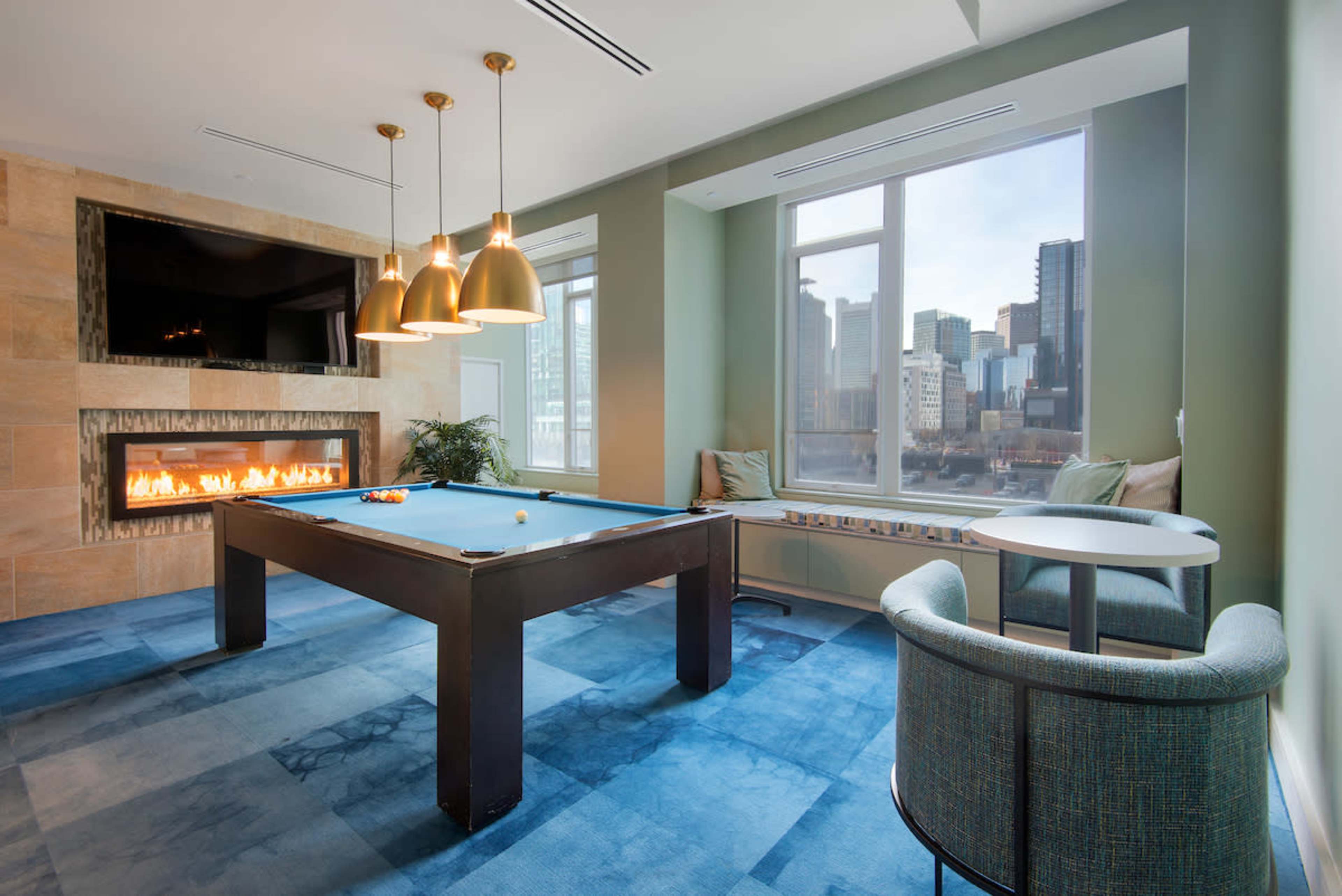 A modern game room featuring a pool table, a wall-mounted television, and large windows with a cityscape view.