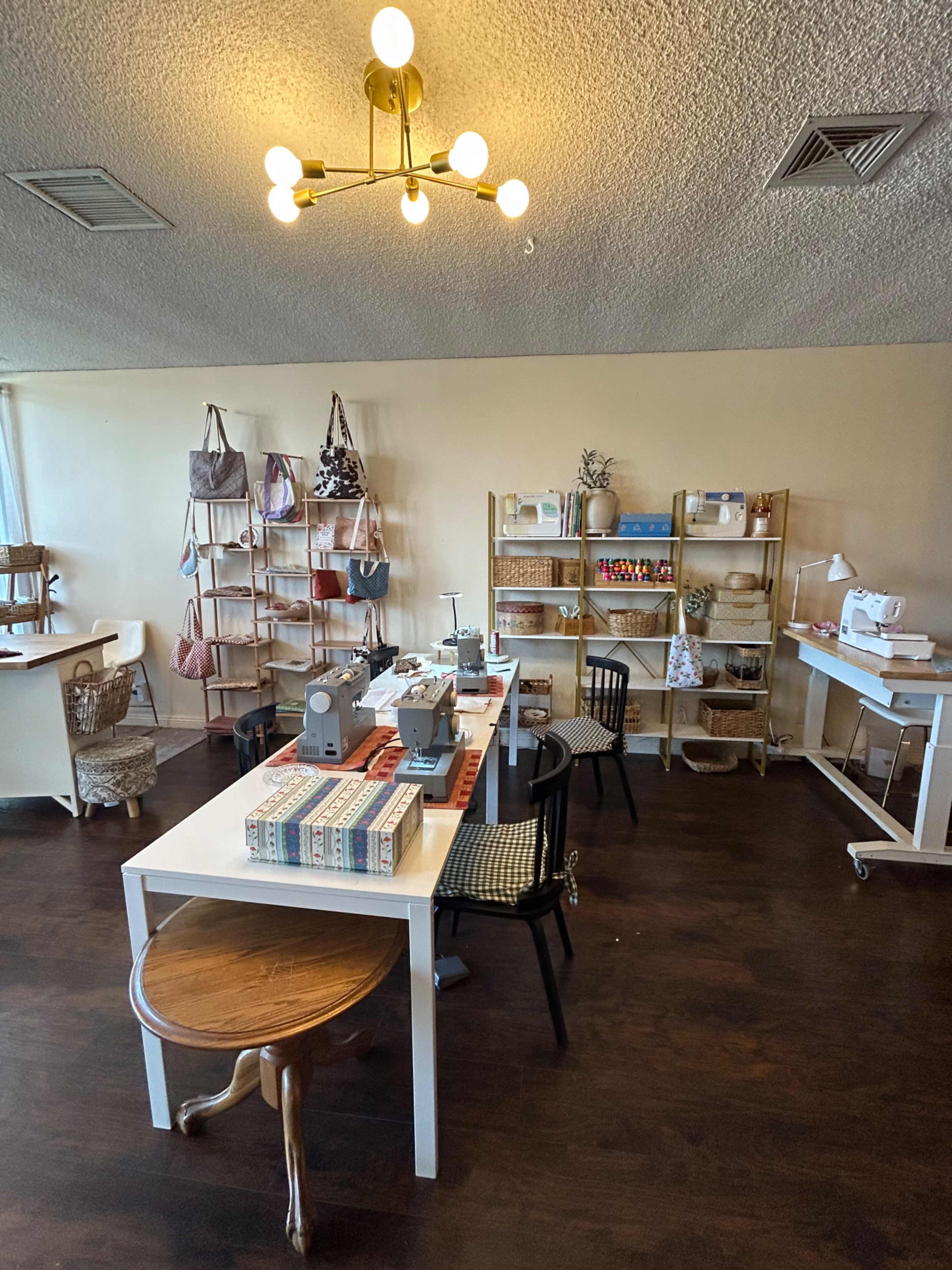 The image shows a sewing studio with wooden shelves displaying craft supplies, a central table with sewing machines, and various chairs arranged around the space.