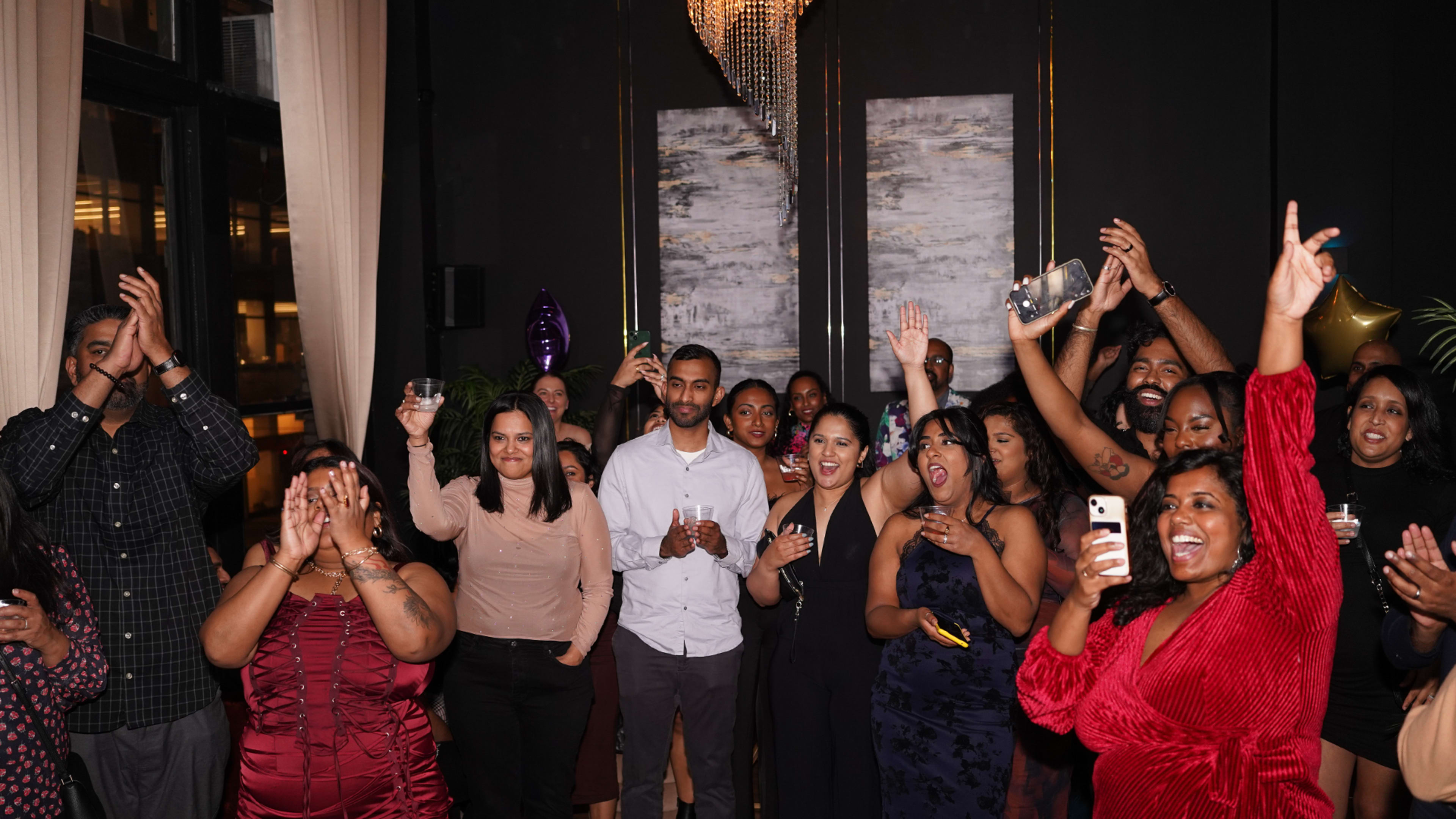 A large group of people is celebrating indoors, with many raising their arms and drinks in a lively atmosphere.