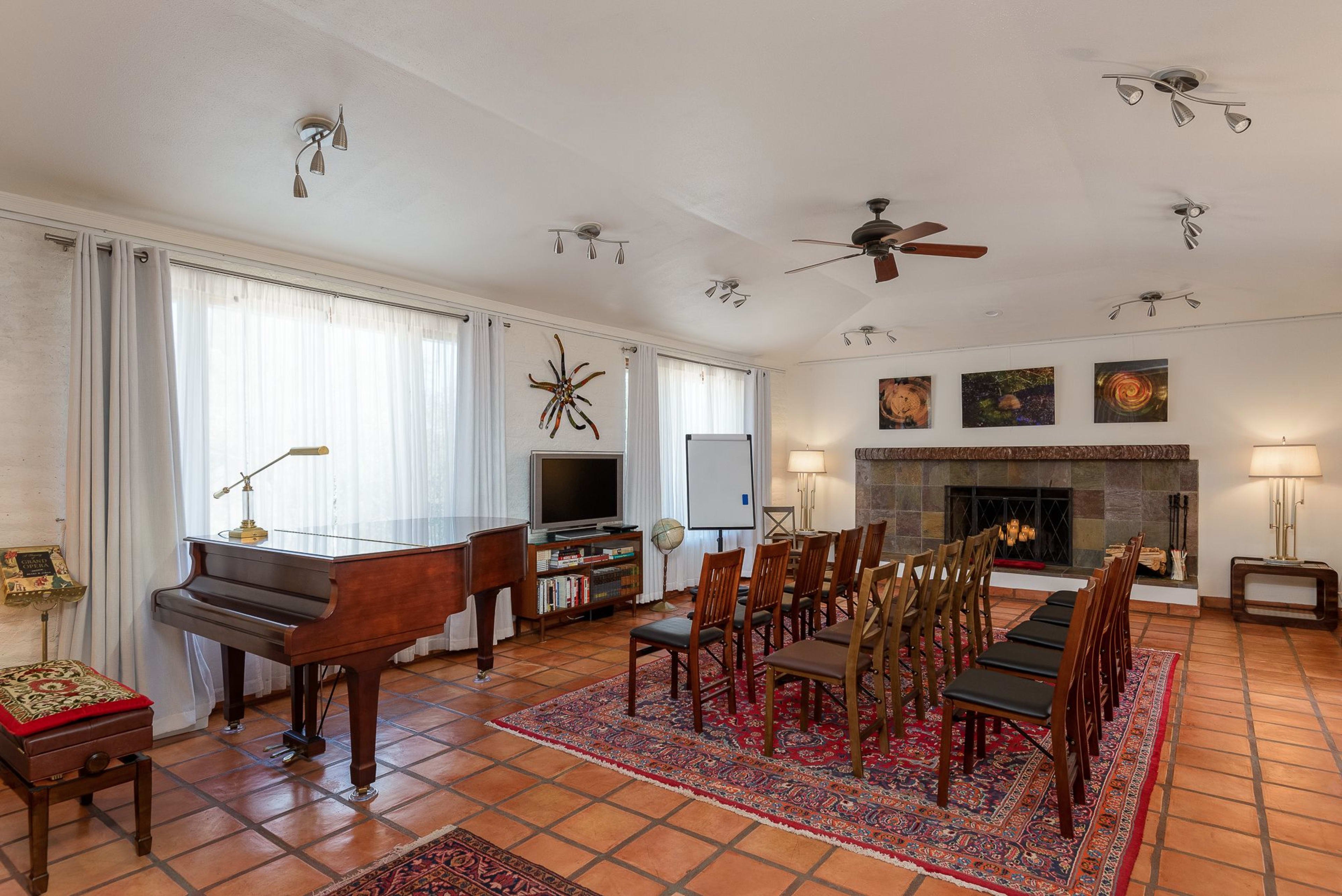 The room features a grand piano, a seating arrangement of wooden chairs, and a fireplace, all surrounded by large windows and decorative wall art.