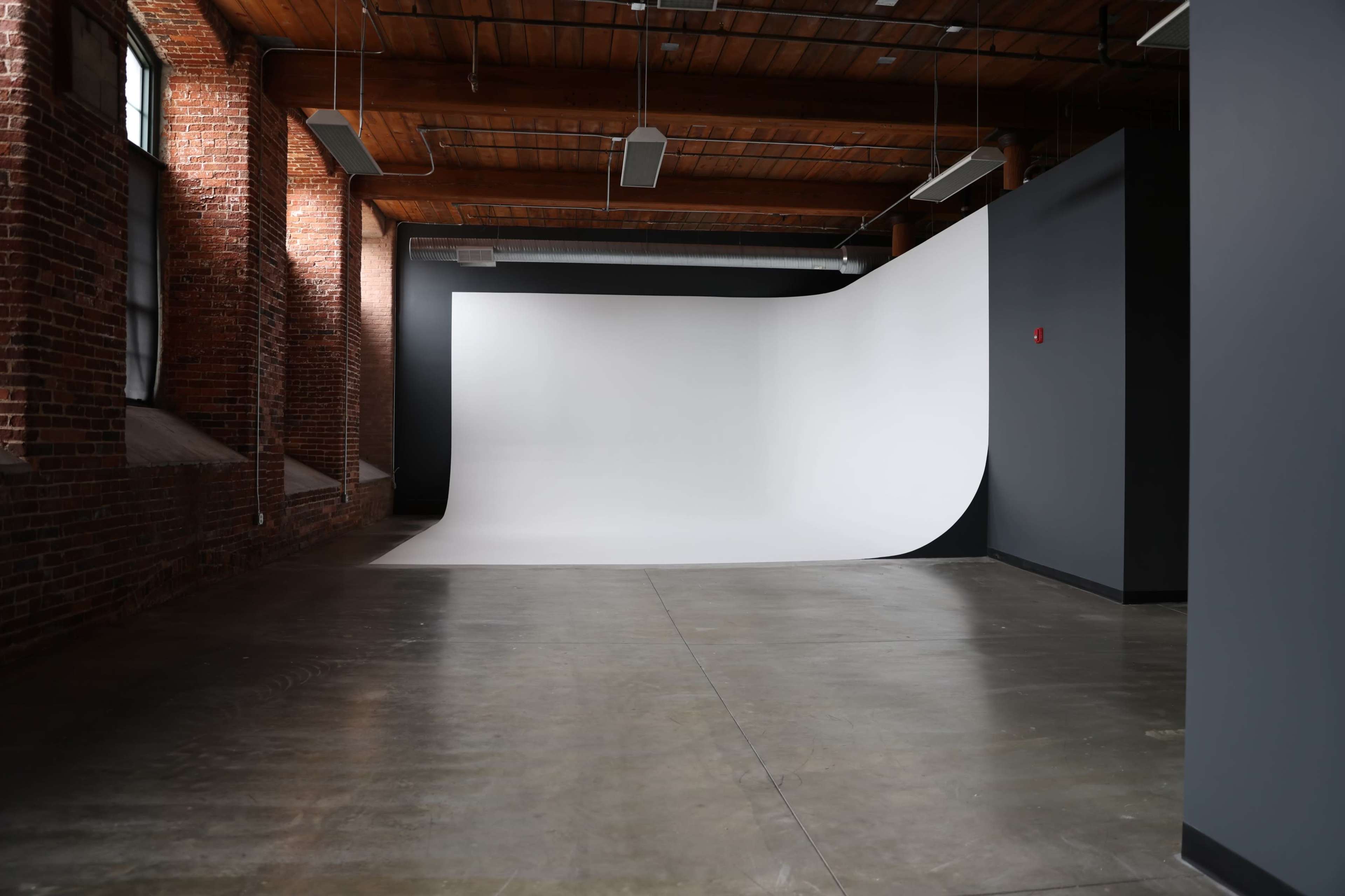 The image shows an empty studio space with brick walls, a wooden ceiling, and a seamless white backdrop extending to the floor.