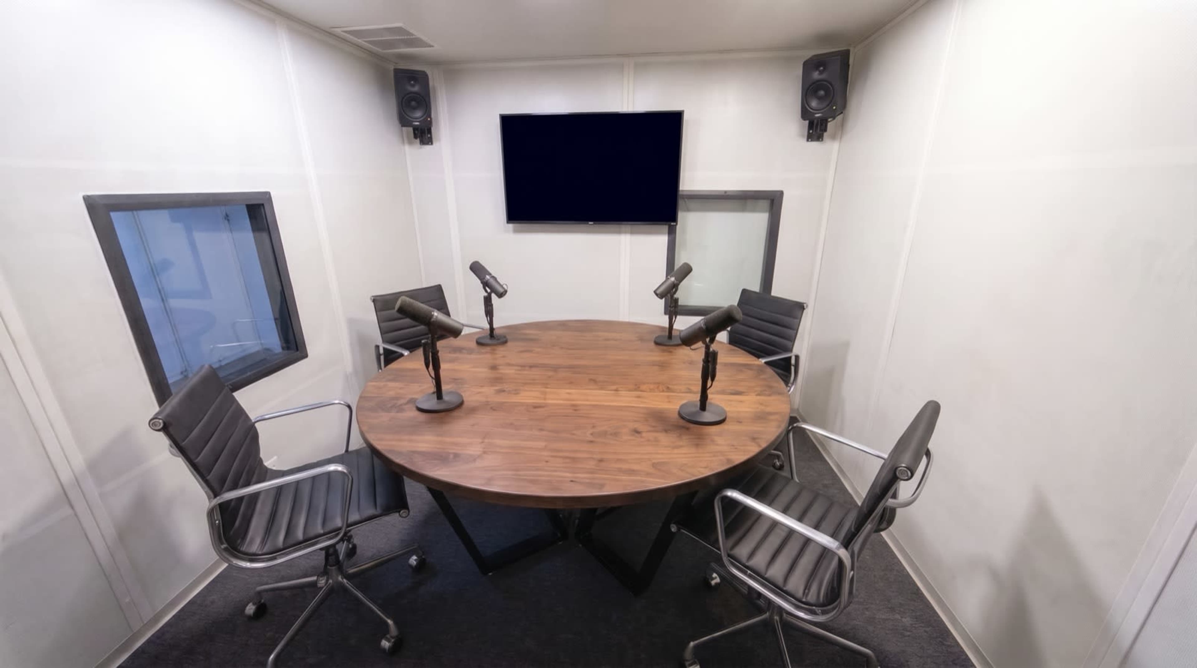 The image shows a small recording studio with a round wooden table, four black chairs, and two microphones arranged around the table, along with a wall-mounted screen and speakers.