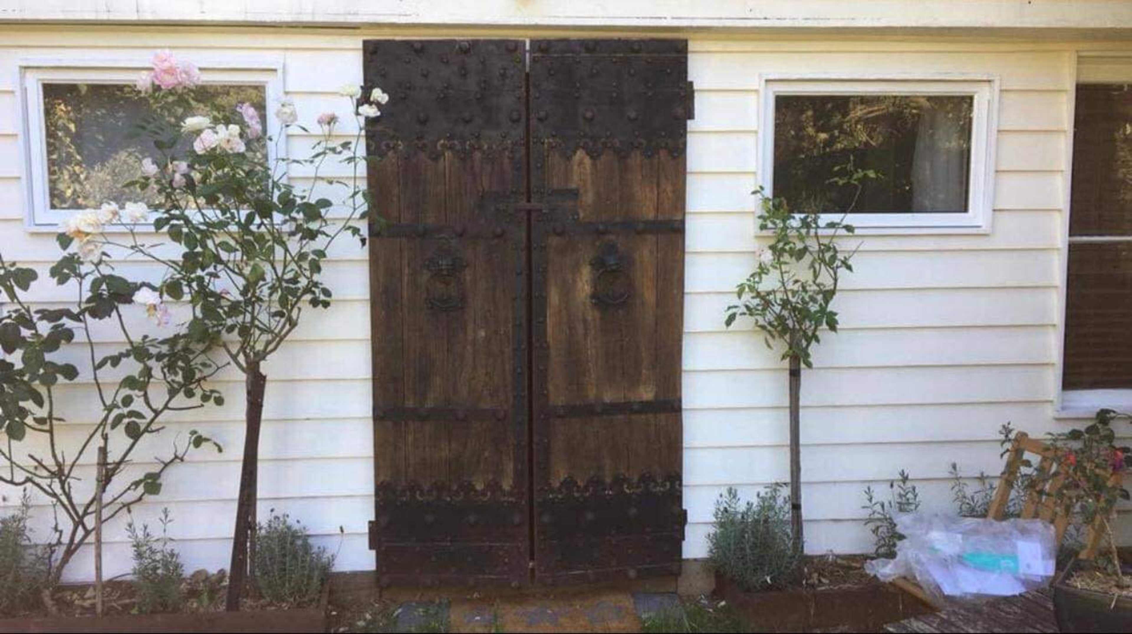 A pair of wooden double doors with metal accents, flanked by two small trees, set against a white house exterior.