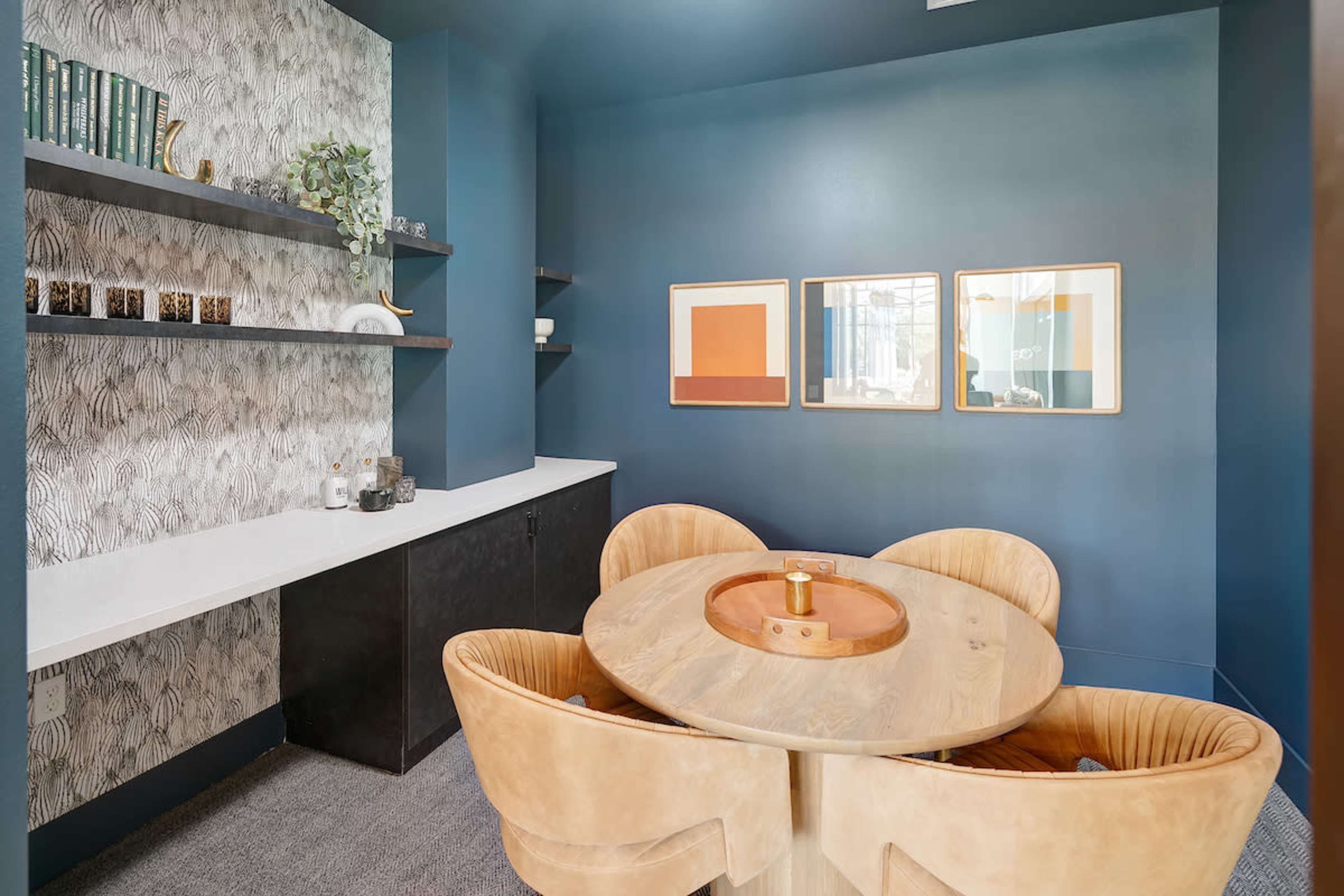 A modern, small meeting room featuring a round wooden table with four tan chairs, framed artwork on the walls, and a minimalist shelf along one side.