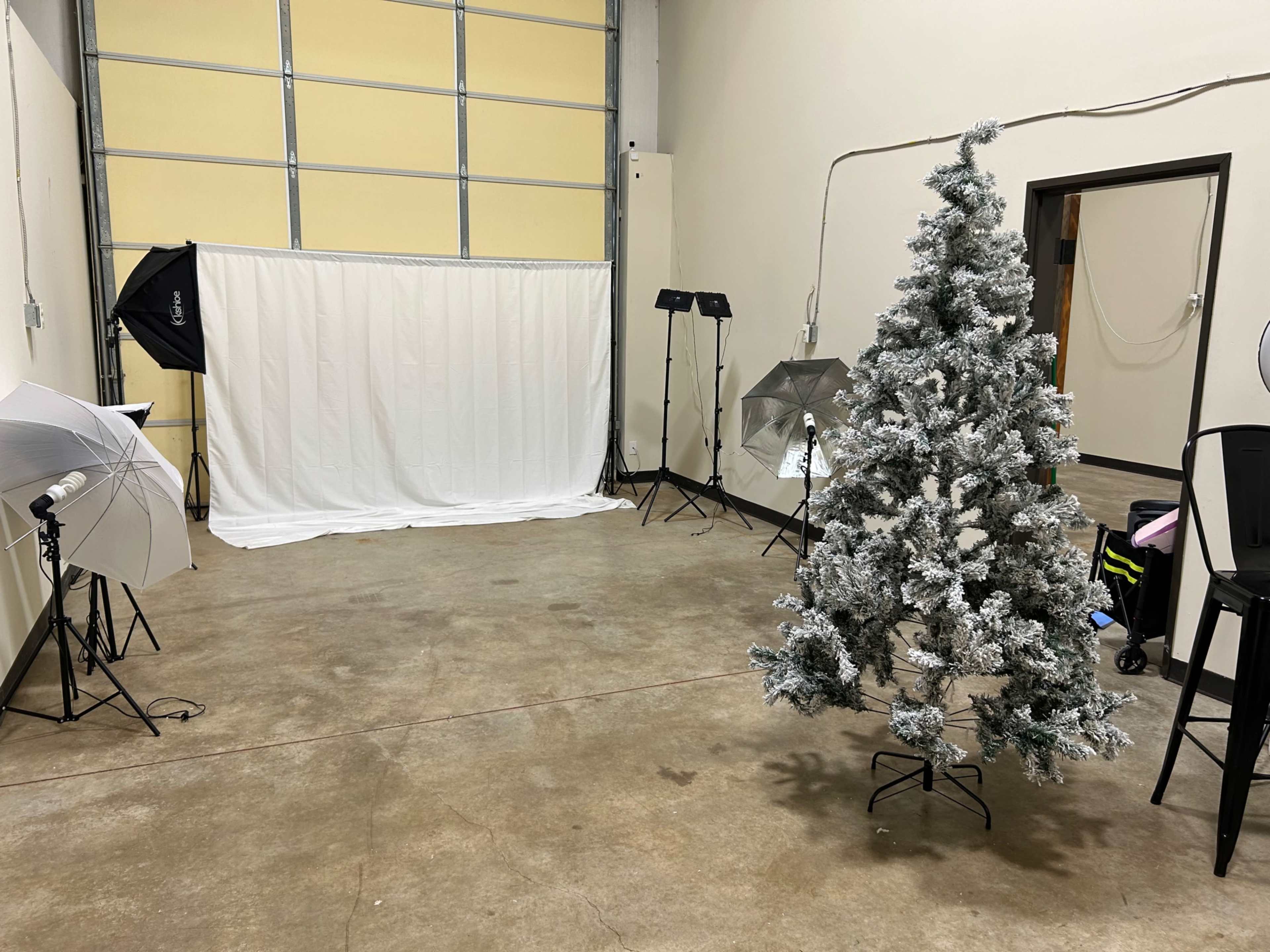 The image shows a spacious indoor area with a frosted artificial Christmas tree, a white backdrop, and several lighting equipment setups.
