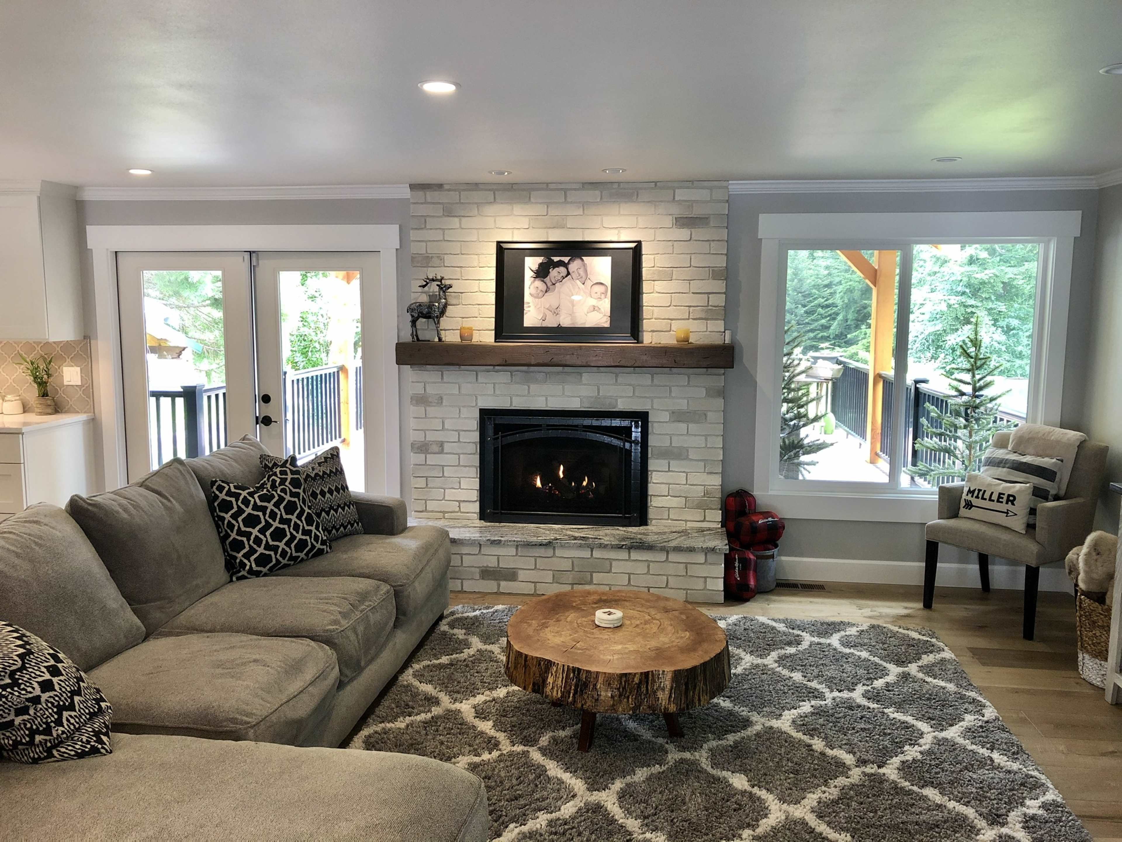 The living room features a gray sectional sofa, a stone fireplace with a framed picture above, a round wooden coffee table, and large windows overlooking a deck.