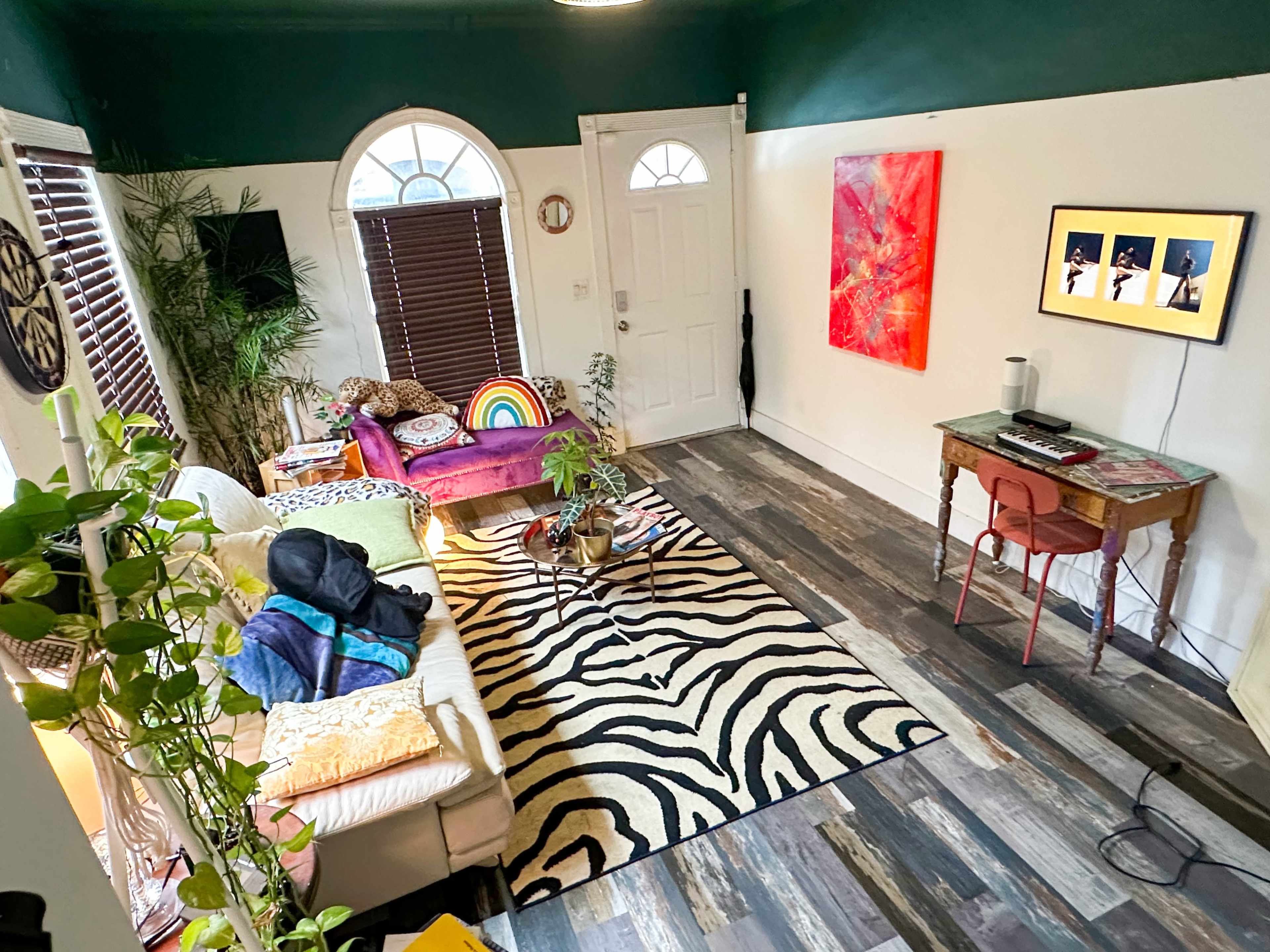 The room features a colorful seating area with a rainbow-striped cushion, a zebra-patterned rug, and a small desk with a monitor and chair against a vibrant green and white wall.