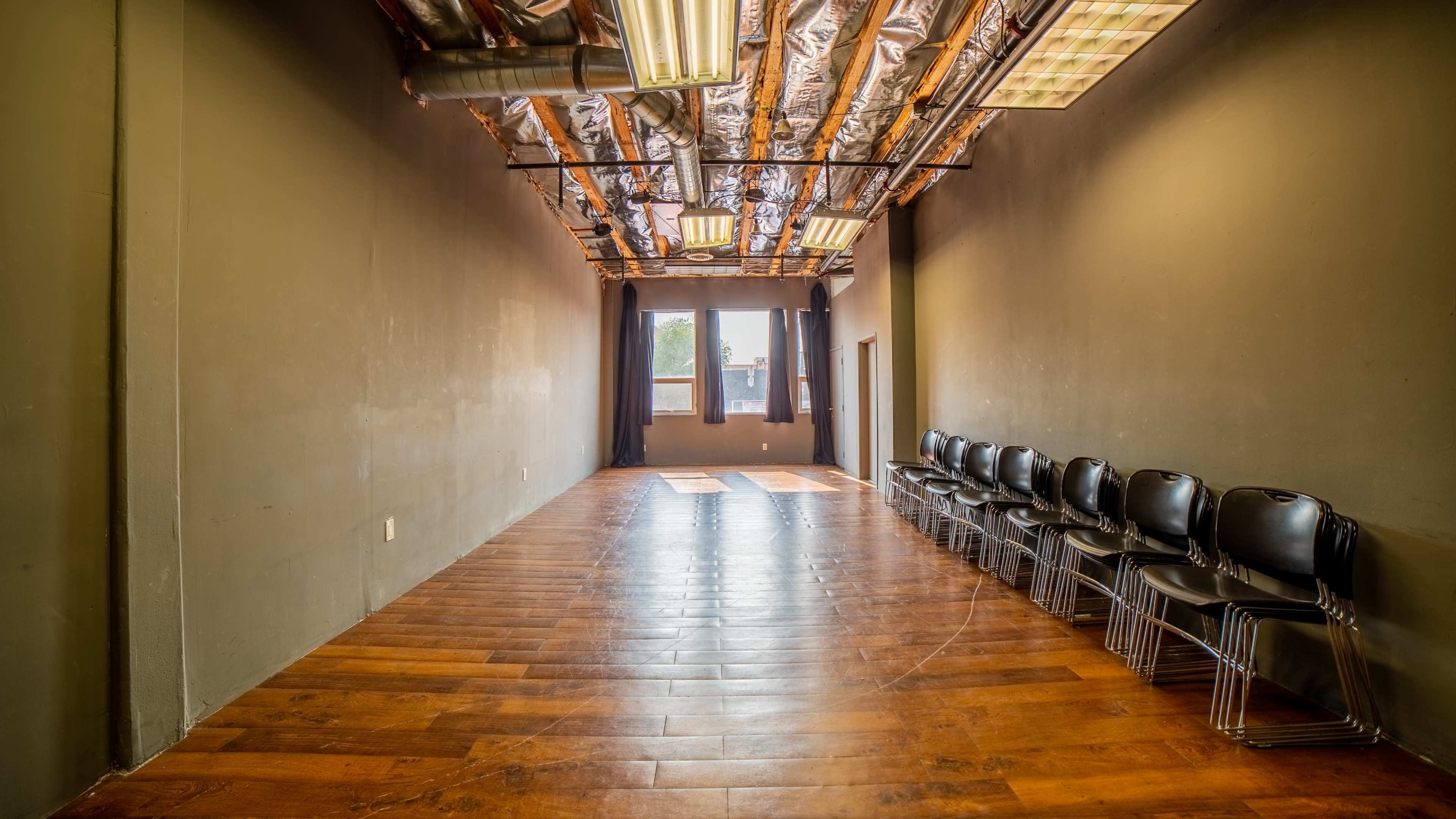 A long, narrow room with wooden flooring and a row of black chairs along one wall, illuminated by natural light from windows at the far end.