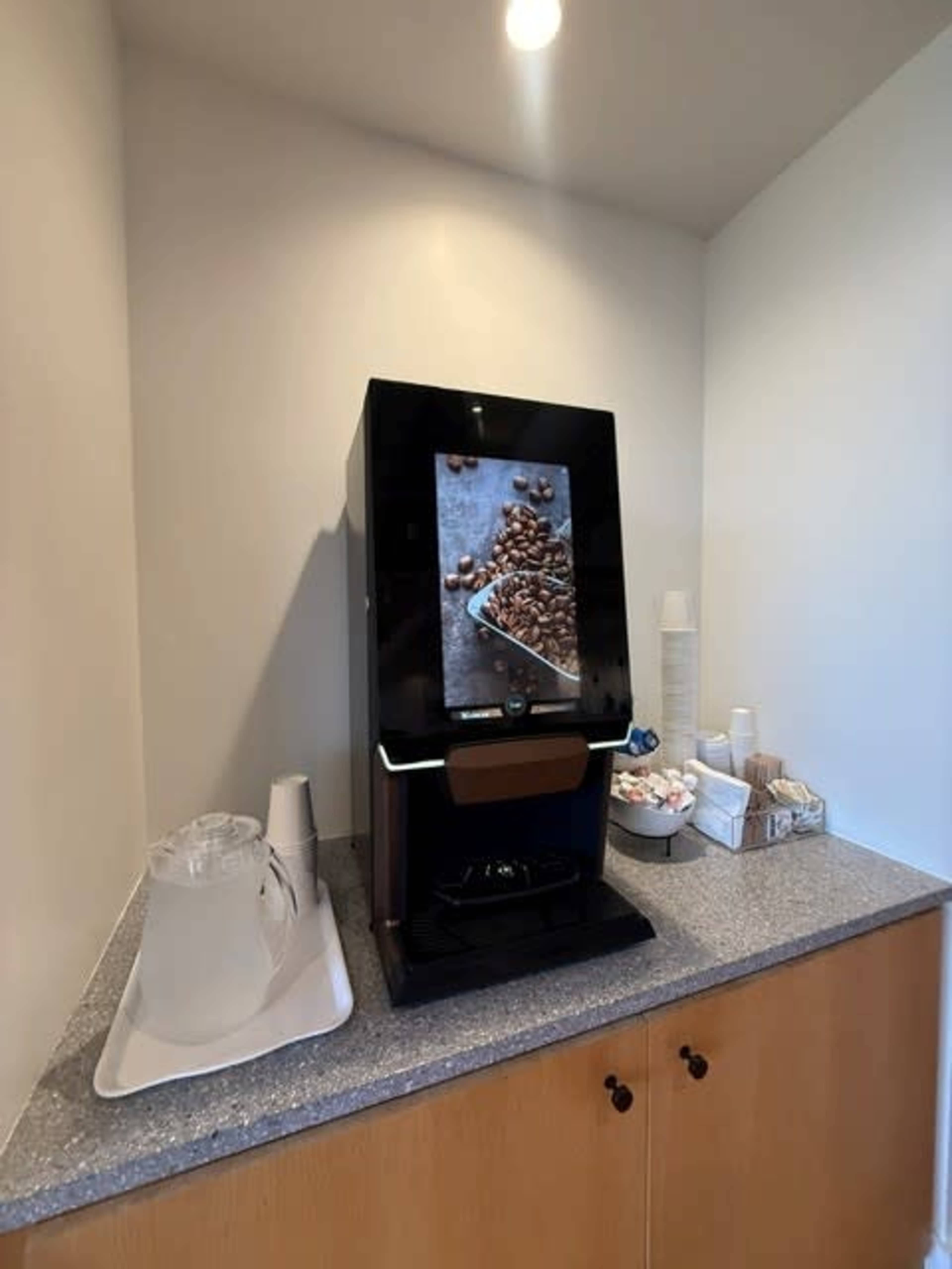 A coffee machine is placed on a gray countertop against a light-colored wall, with a pitcher, cups, and sugar packets nearby.