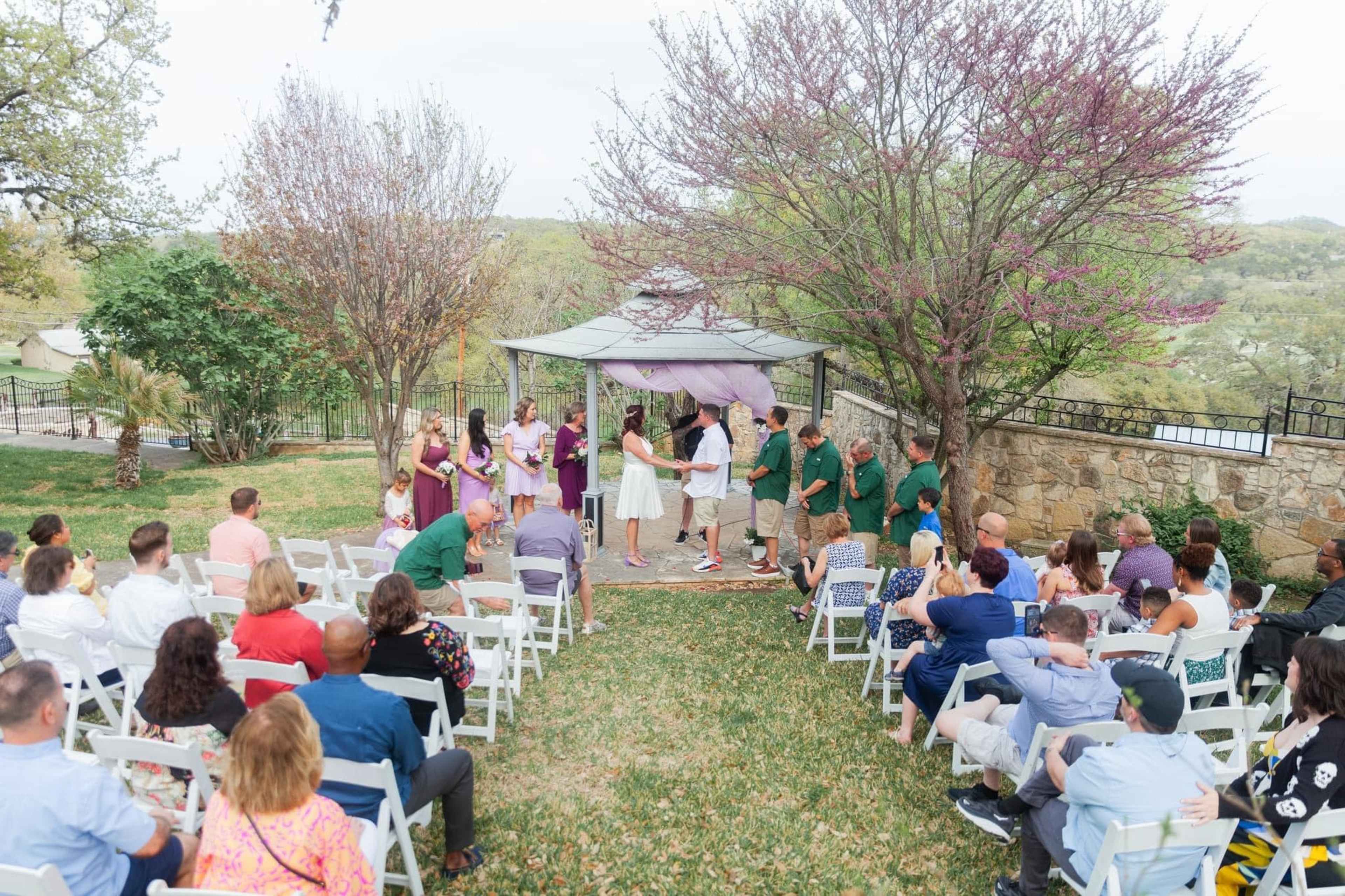 A wedding ceremony is taking place outdoors under a decorative canopy, surrounded by guests seated on white chairs.