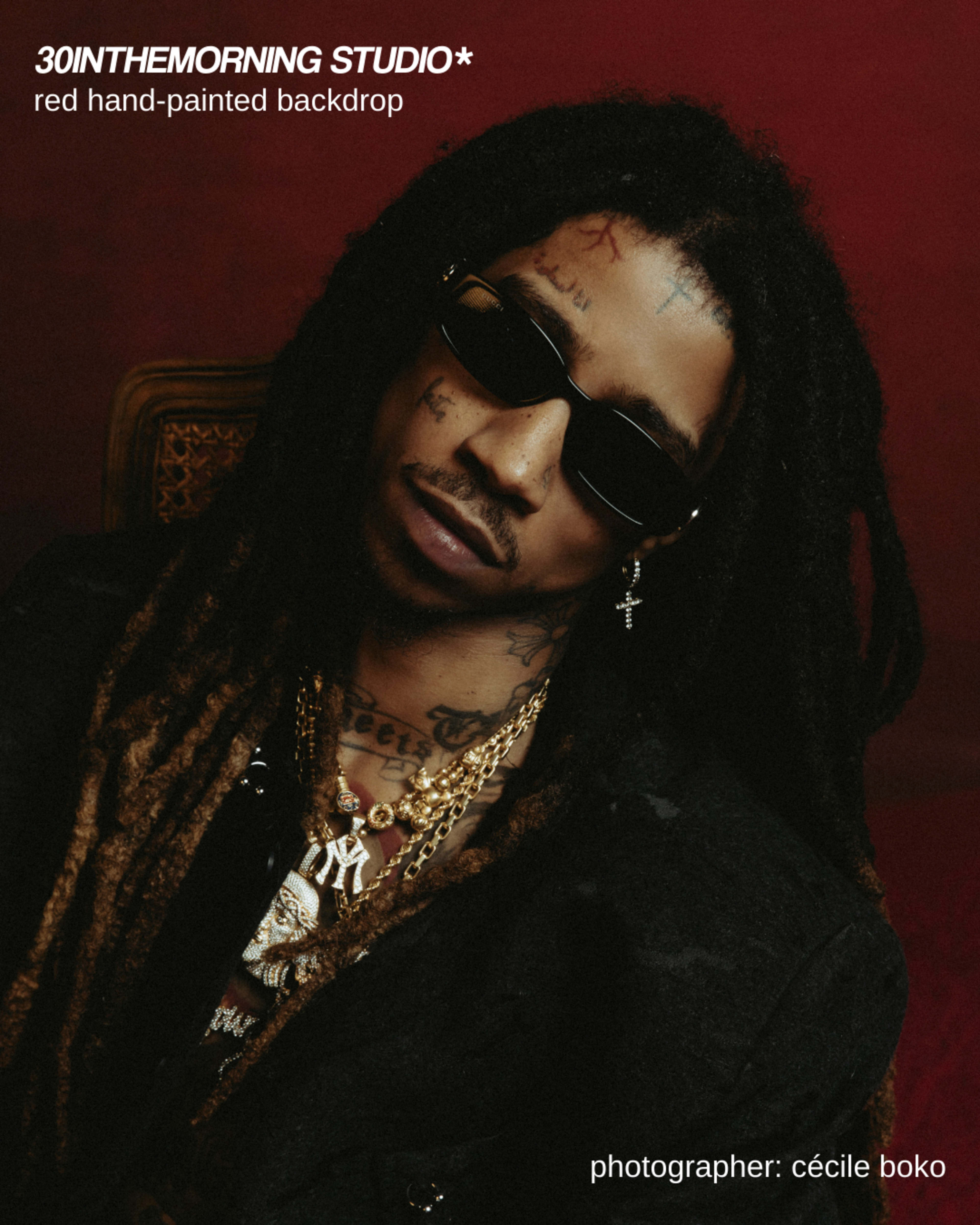 The image shows a person with long, dreadlocked hair wearing sunglasses and multiple gold chains, posed against a red hand-painted backdrop.