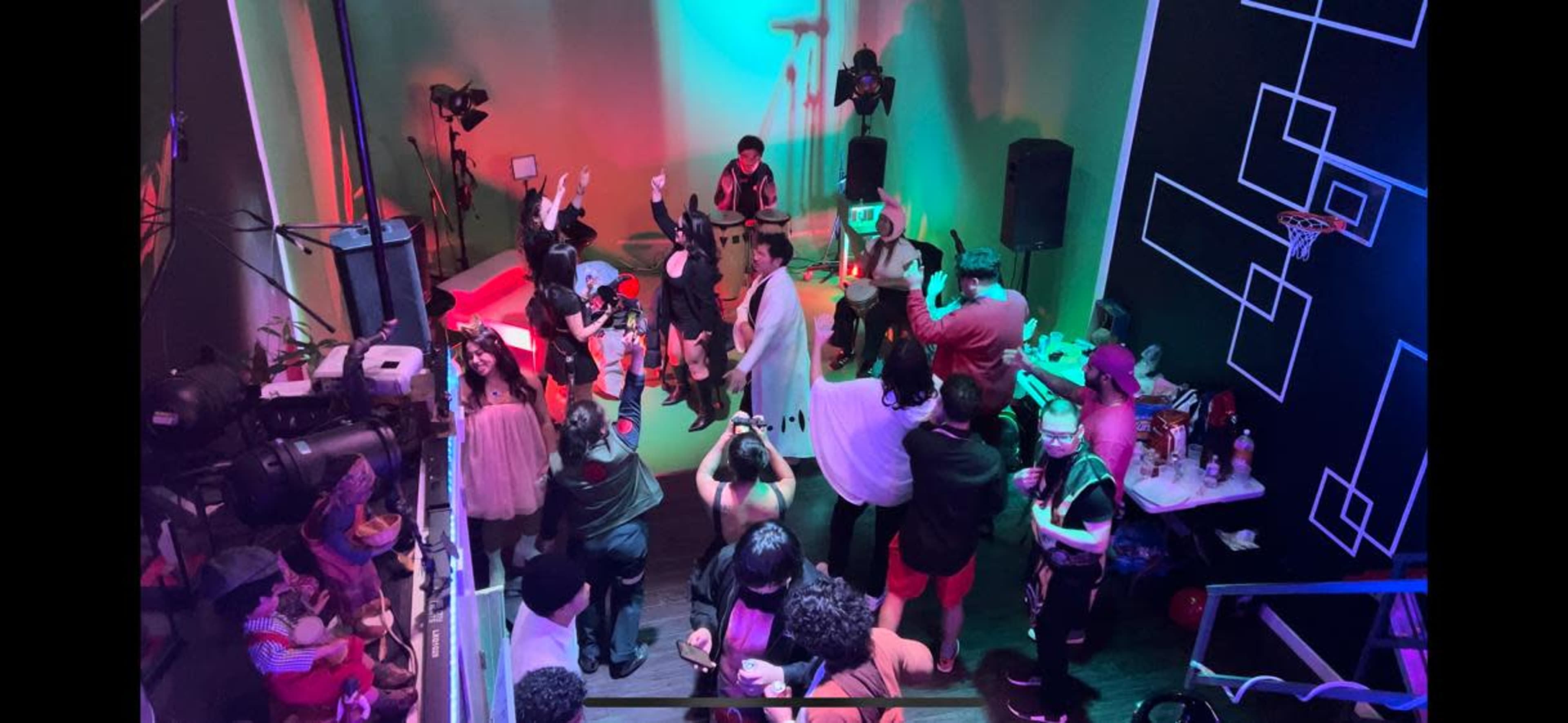 A large group of people gathers in a vibrant, dimly lit venue for a live performance, with cameras and lighting equipment positioned around the room.