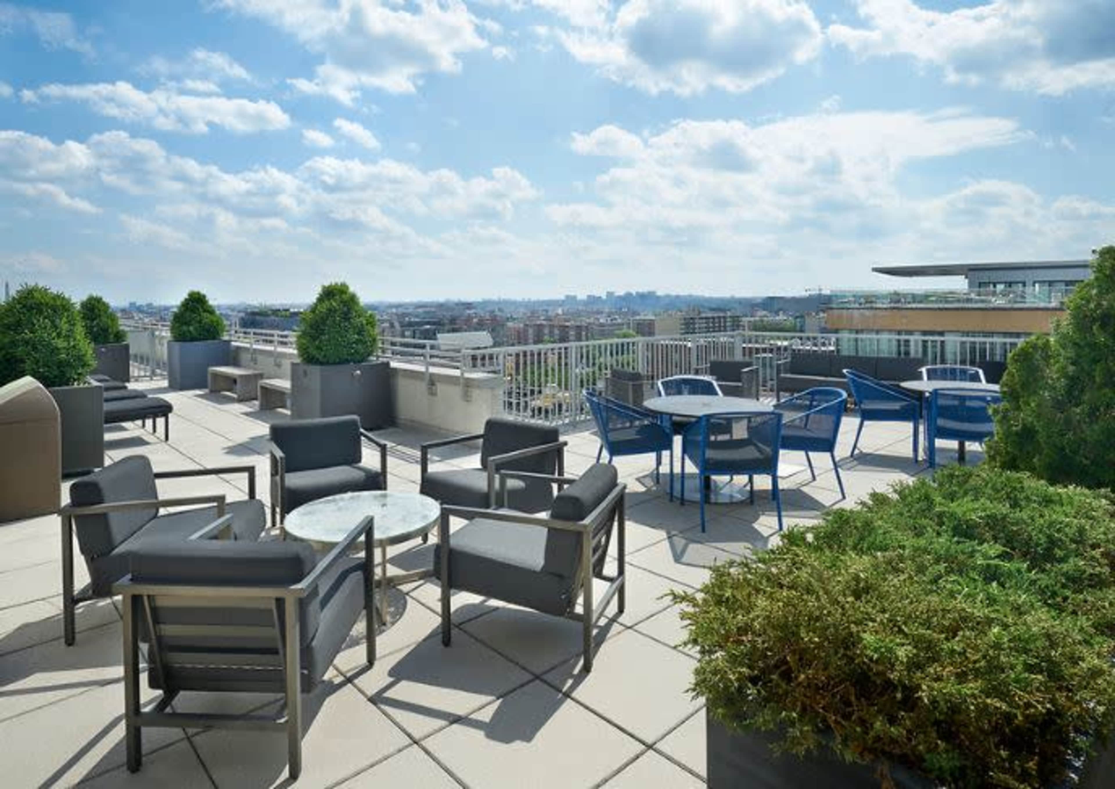 A rooftop terrace with seating arrangements, including chairs and tables, overlooking a city skyline under a partly cloudy sky.