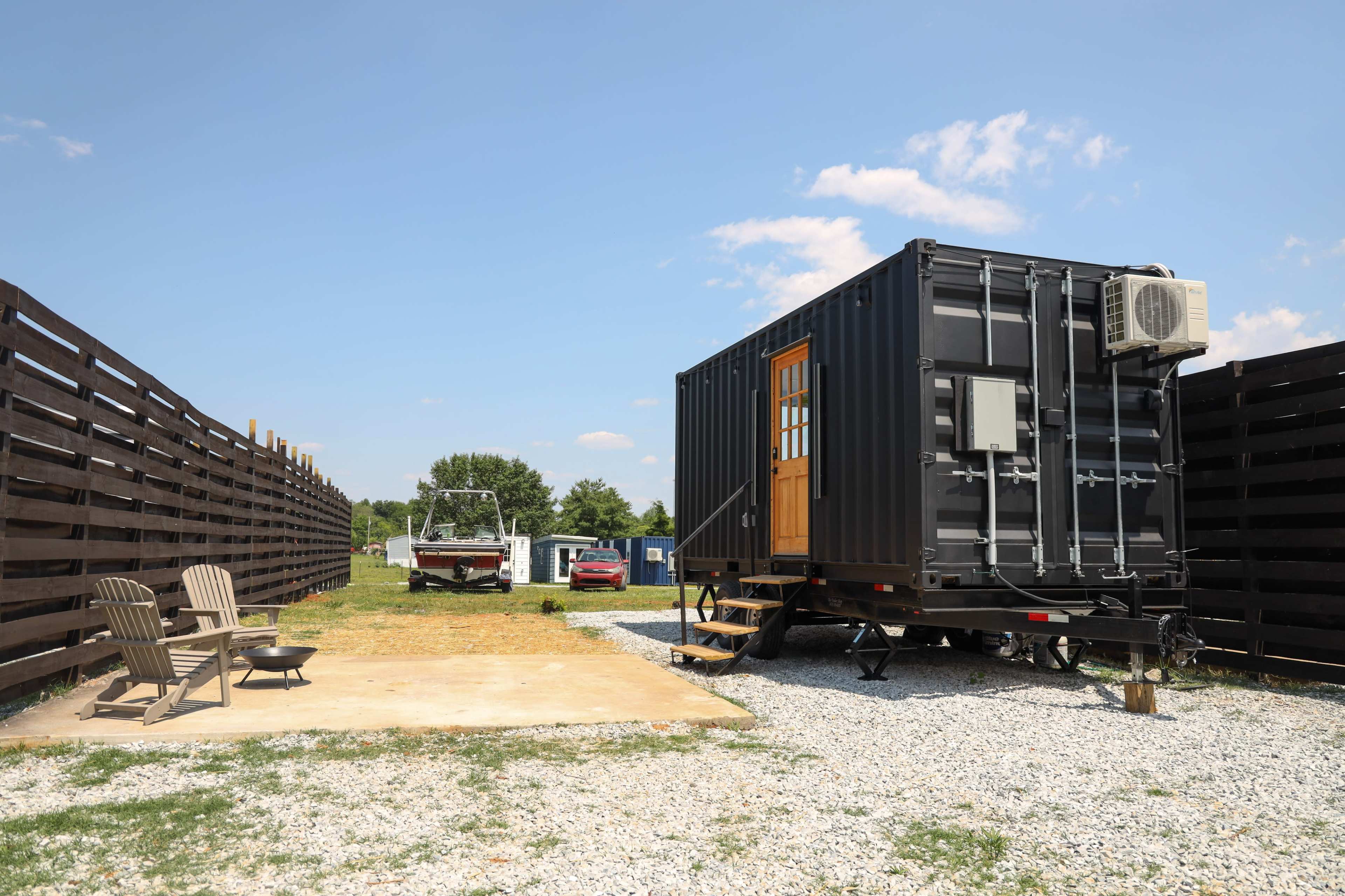 A black shipping container converted into a tiny house is parked next to a gravel area with seating and a fire pit, surrounded by wooden fencing.
