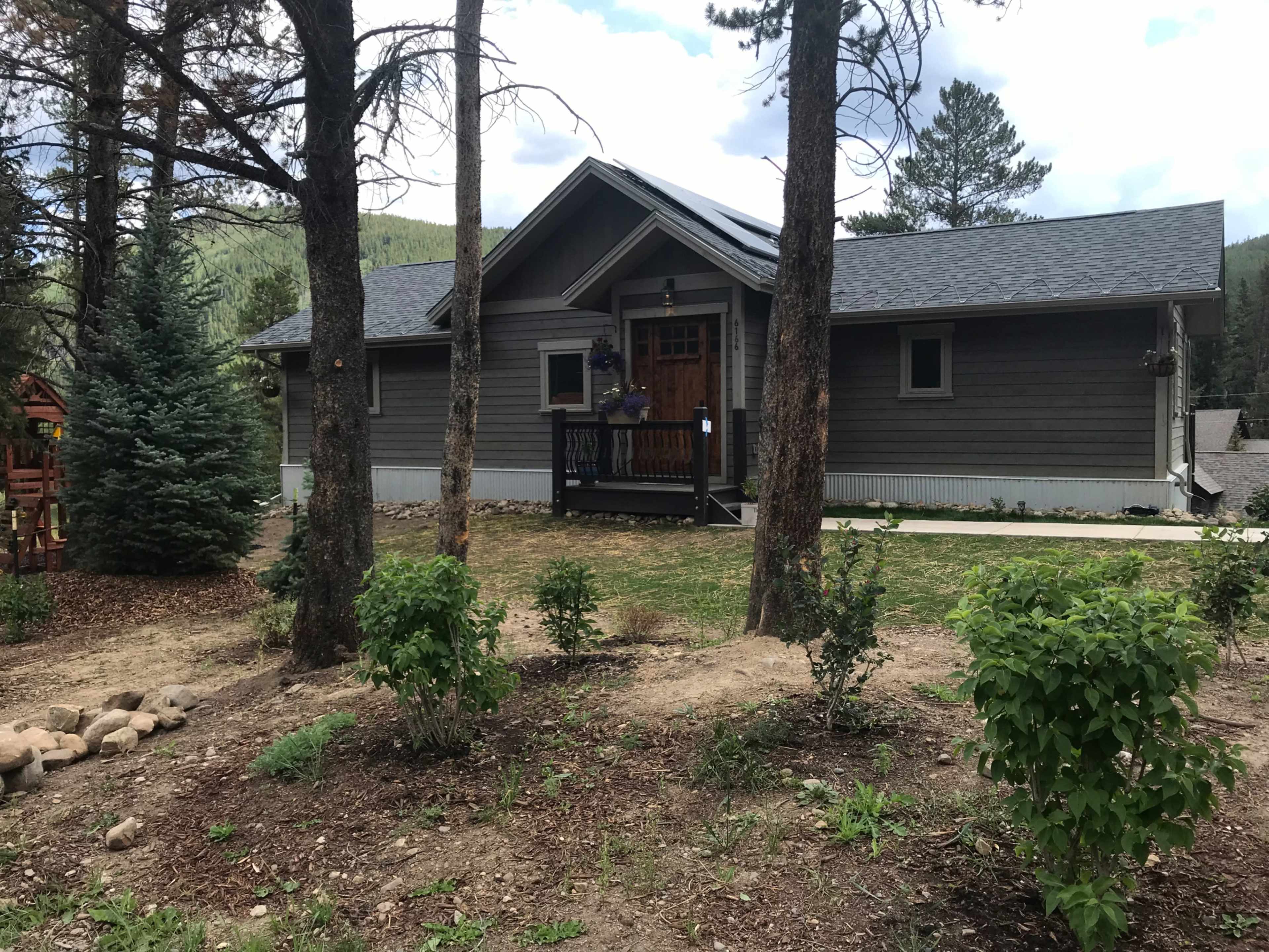 A gray wooden house stands among tall trees, with a front porch and landscaped yard.