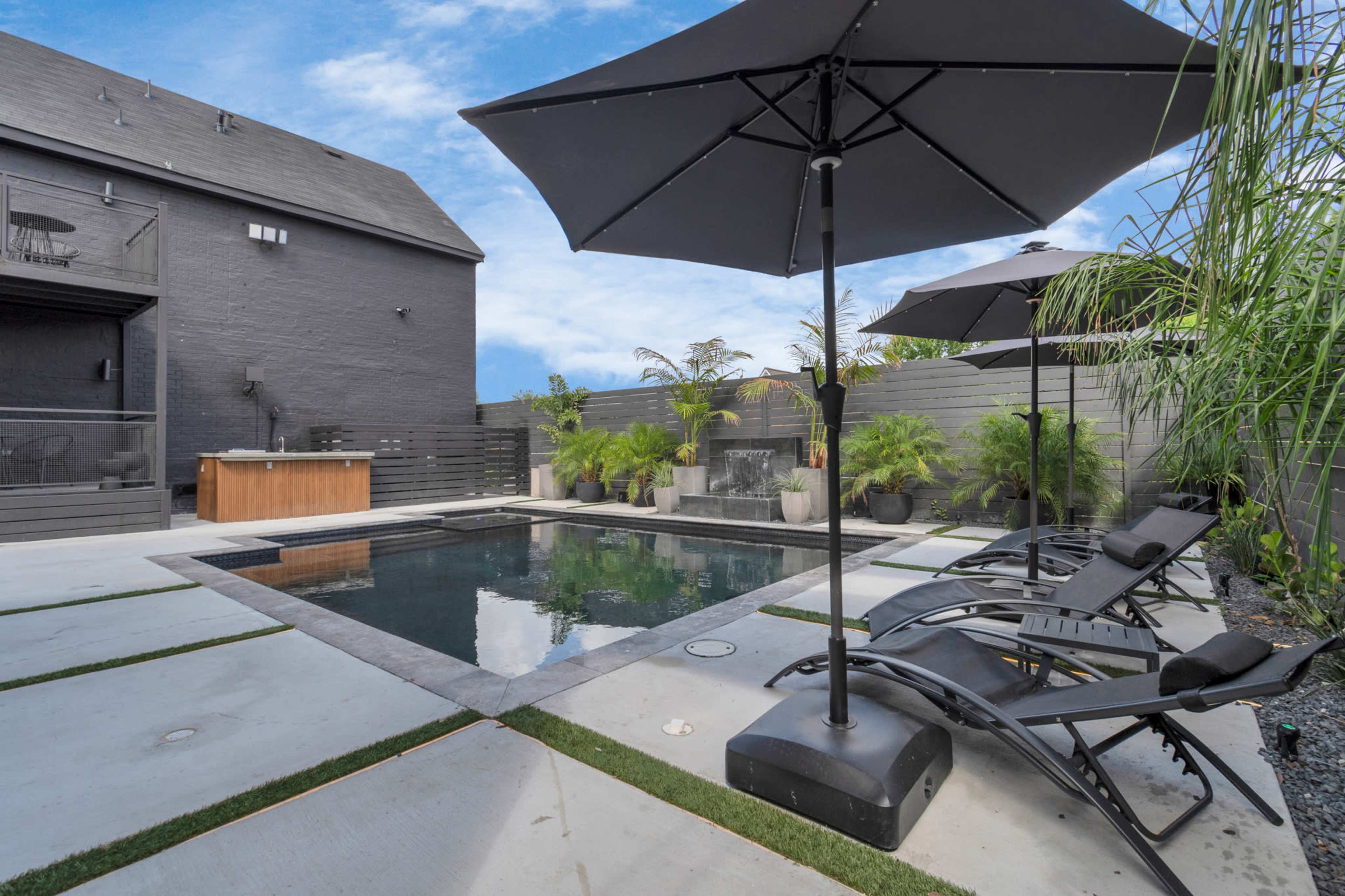 The image shows a modern backyard with a rectangular swimming pool surrounded by concrete slabs, lounge chairs, and tall plants, all set against a dark-colored house.