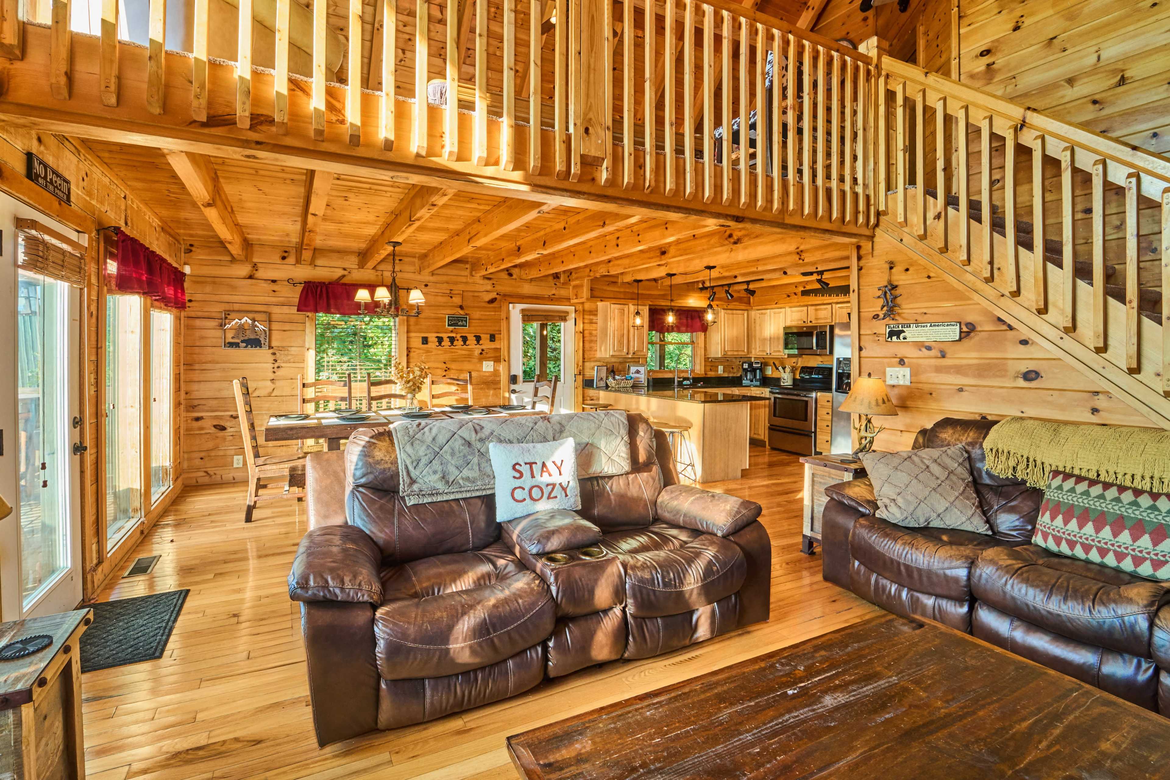 The image shows a wooden cabin interior featuring a living room with leather sofas, a wooden dining table, and a staircase leading to a loft area.
