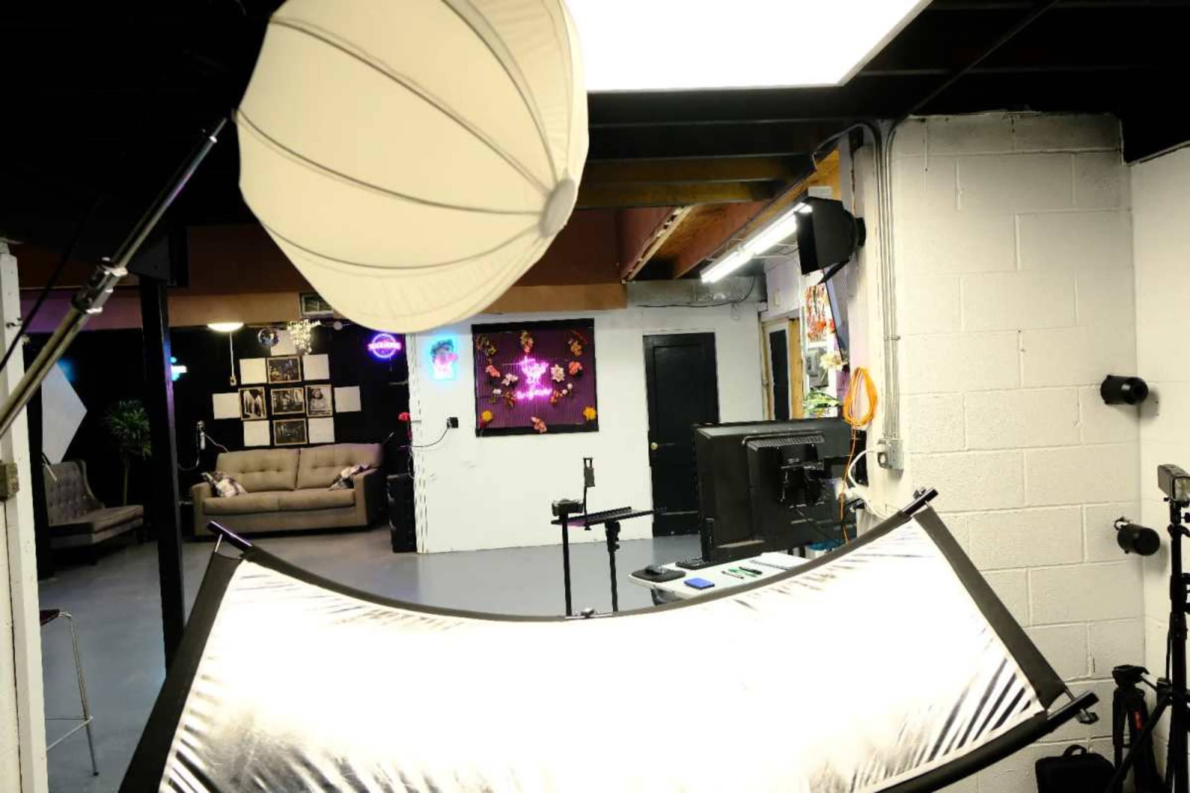 The image shows a photography studio setup featuring lighting equipment, a large reflector, and a backdrop with a couch and artwork visible in the background.