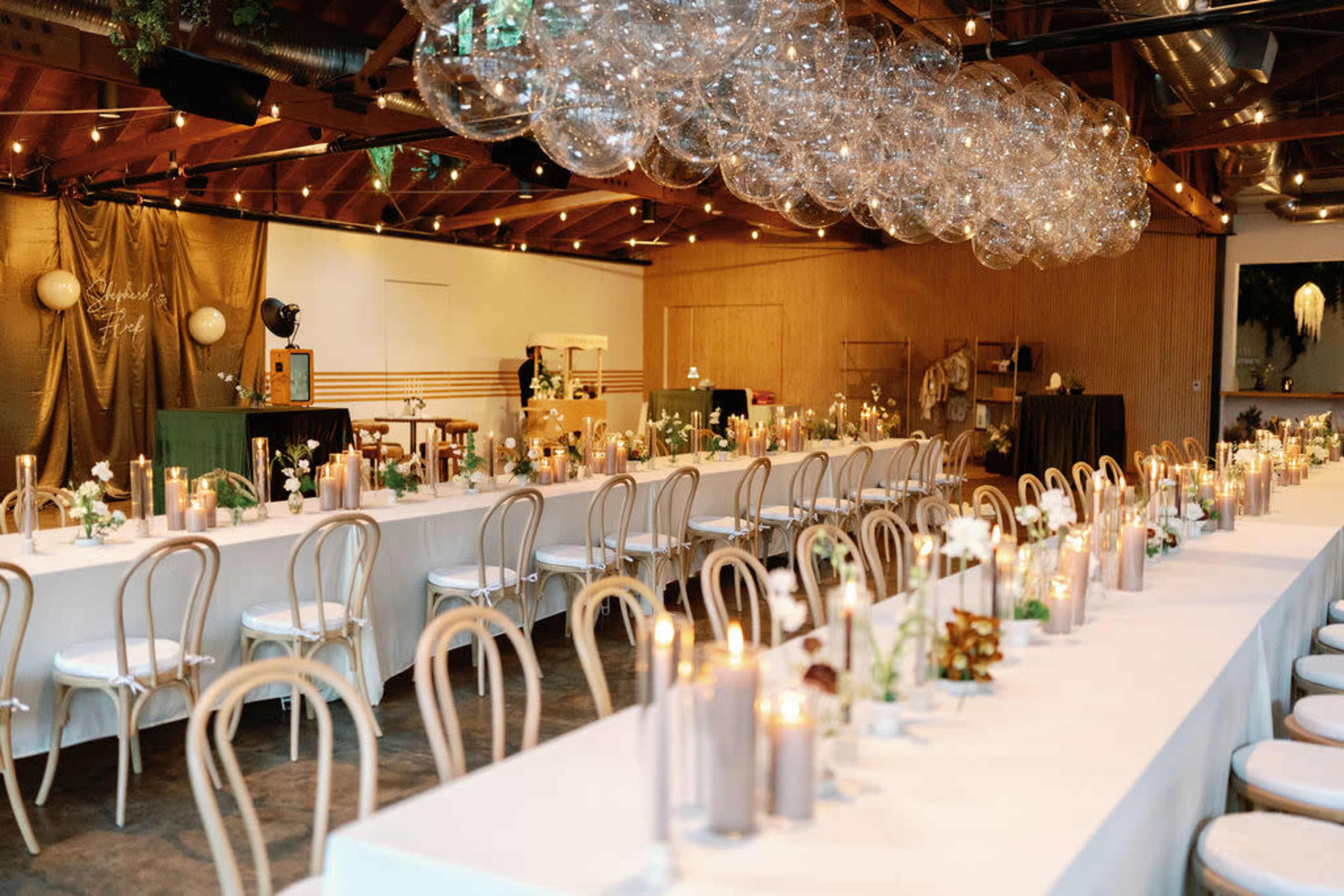 A spacious event venue features long tables decorated with candles and floral centerpieces, beneath a display of hanging glass orbs.