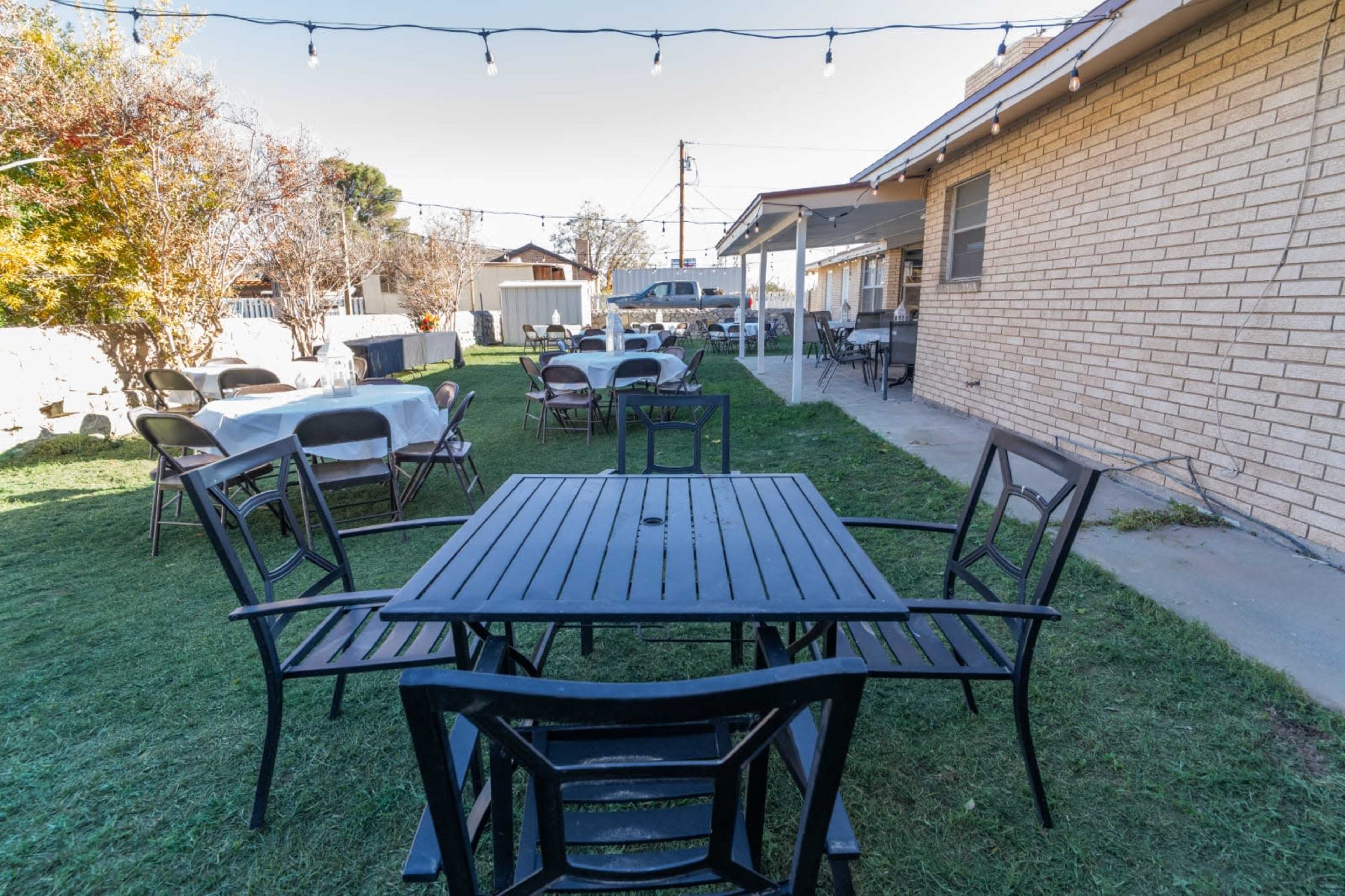 The image shows a backyard outdoor seating area with black metal tables and chairs, surrounded by green grass and string lights overhead.