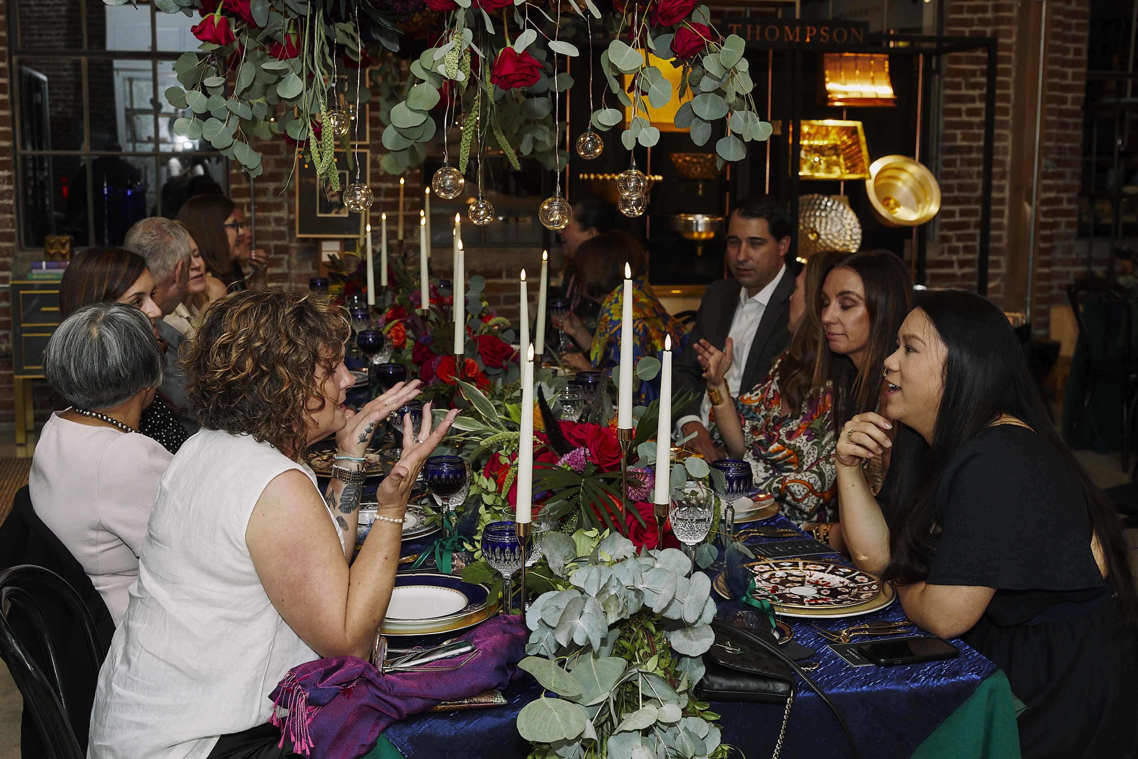 A group of people is gathered around a decorated dining table with candles and floral arrangements, engaged in conversation.