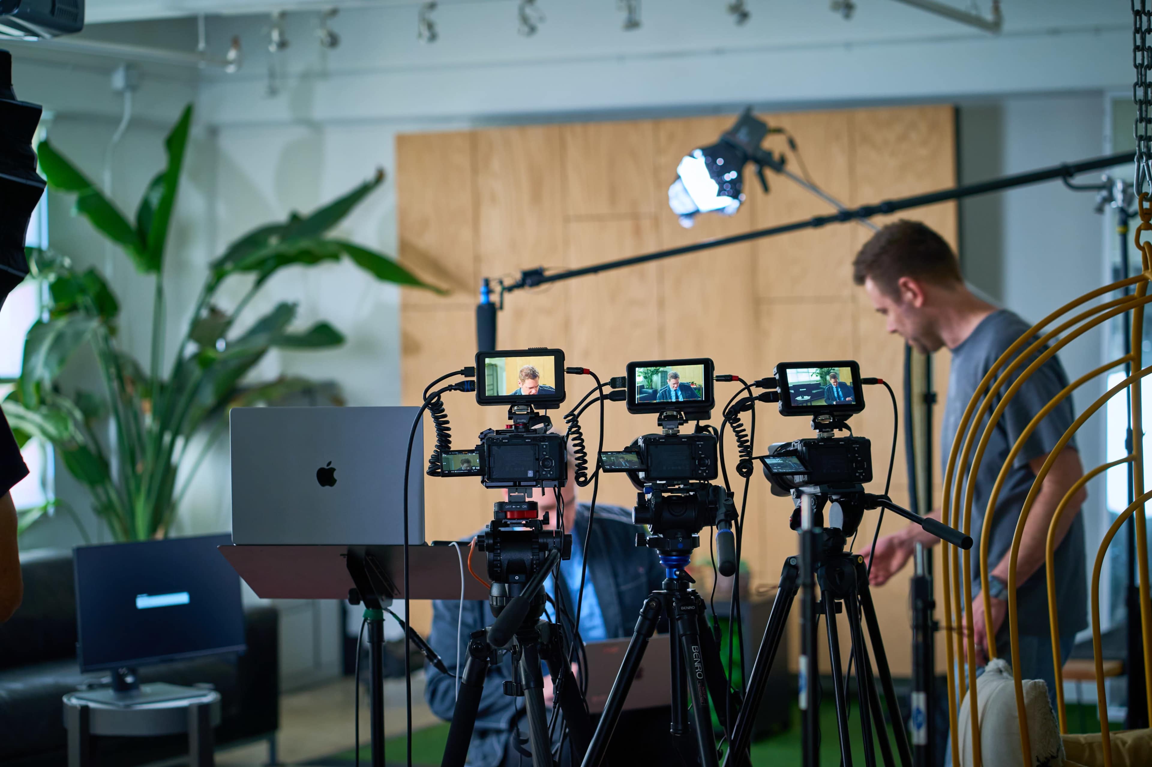A video recording setup features multiple cameras on tripods, with a man adjusting equipment in a well-lit studio space.