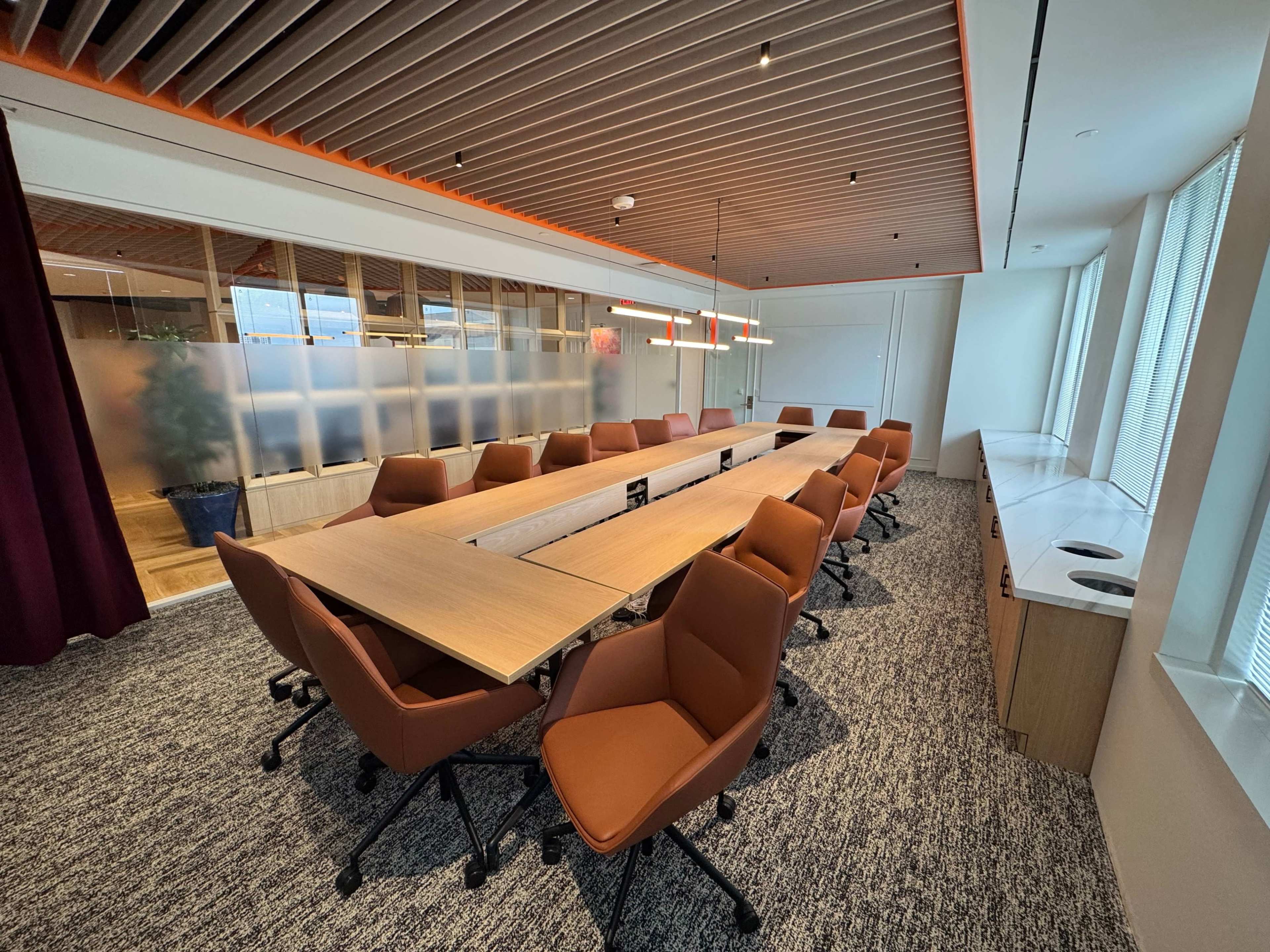 A long conference table surrounded by brown chairs is set up in a well-lit meeting room with large windows.