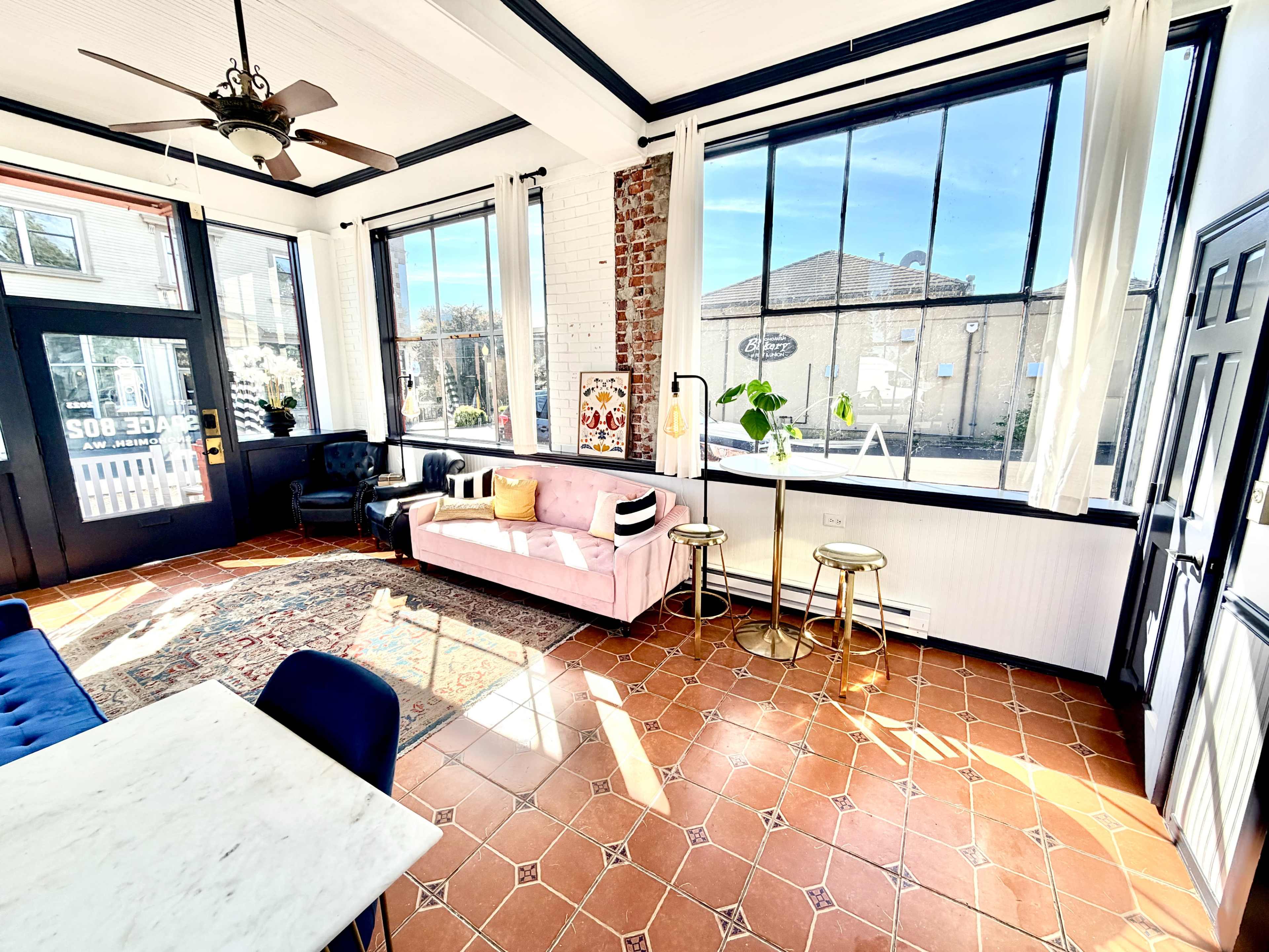 The image shows a bright, sunlit room featuring a pink sofa, two gold side tables, a marble dining table, and large windows with a view of an outdoor area.