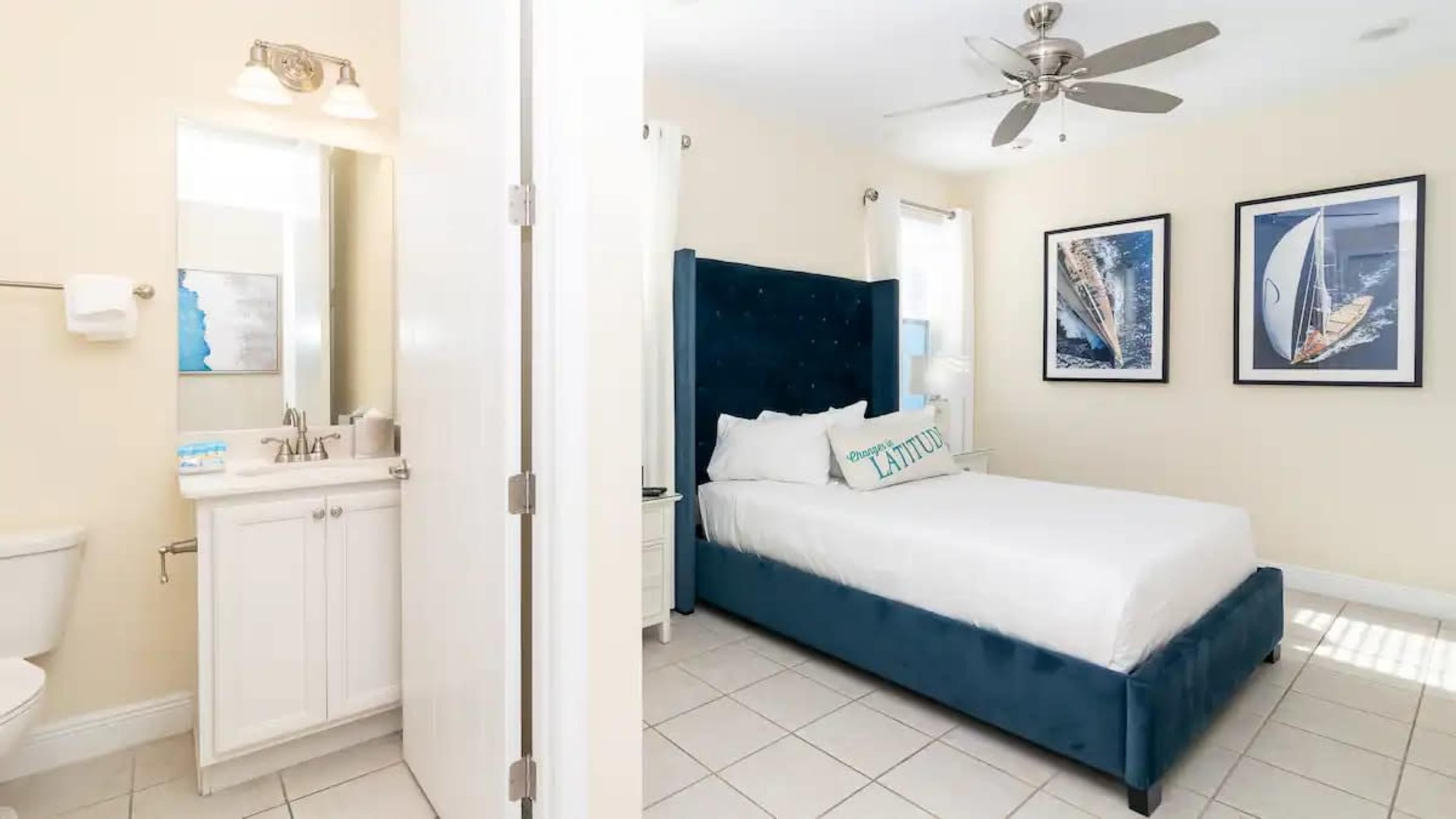 A small, bright bedroom featuring a double bed with a blue upholstered headboard, a nightstand, and decorated with nautical-themed artwork on the walls.