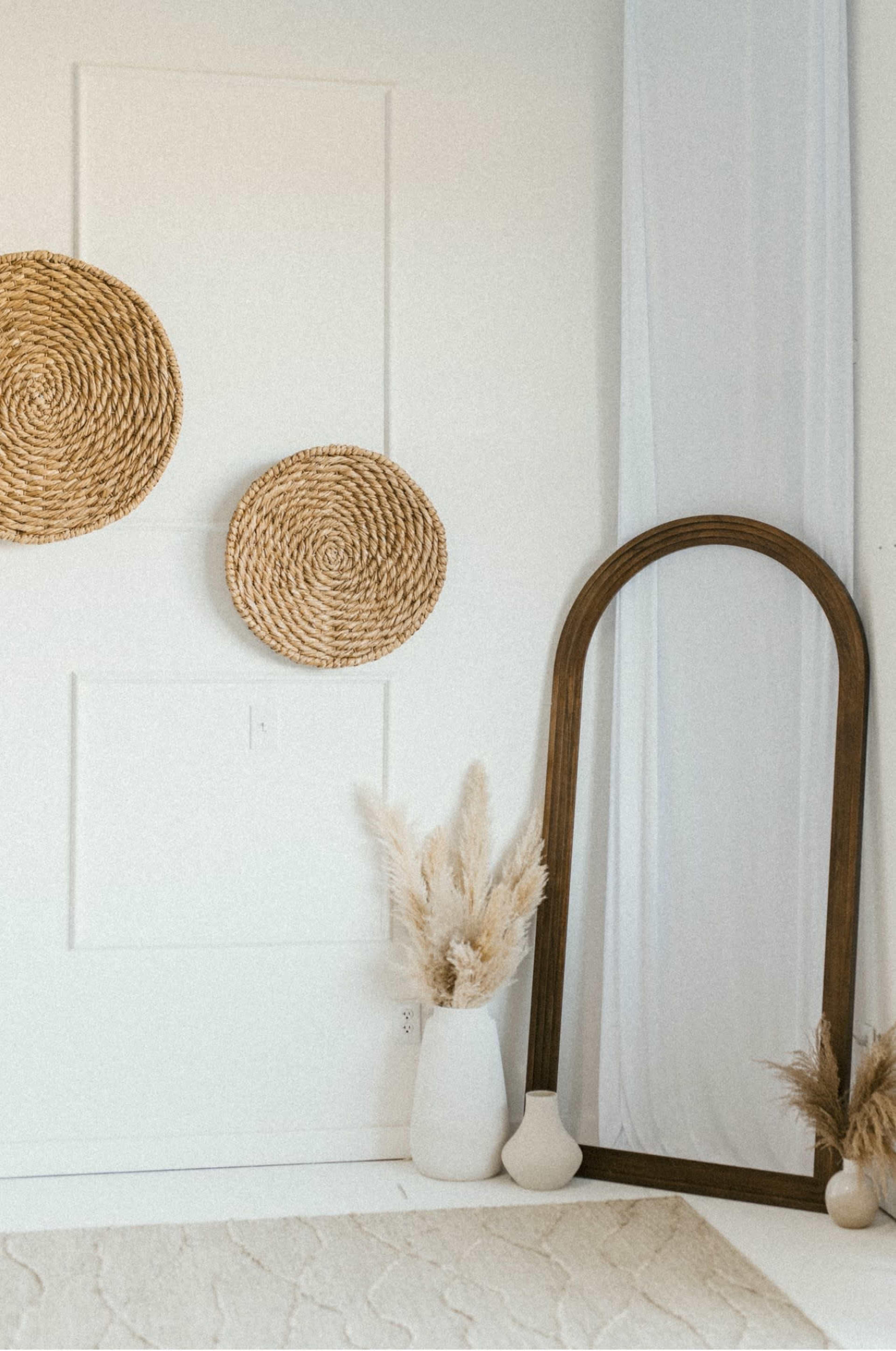 The image features a minimalist interior with two woven decorative wall hangings, a large arched mirror, and a white vase containing fluffy pampas grass, all set against a light-colored wall and floor.