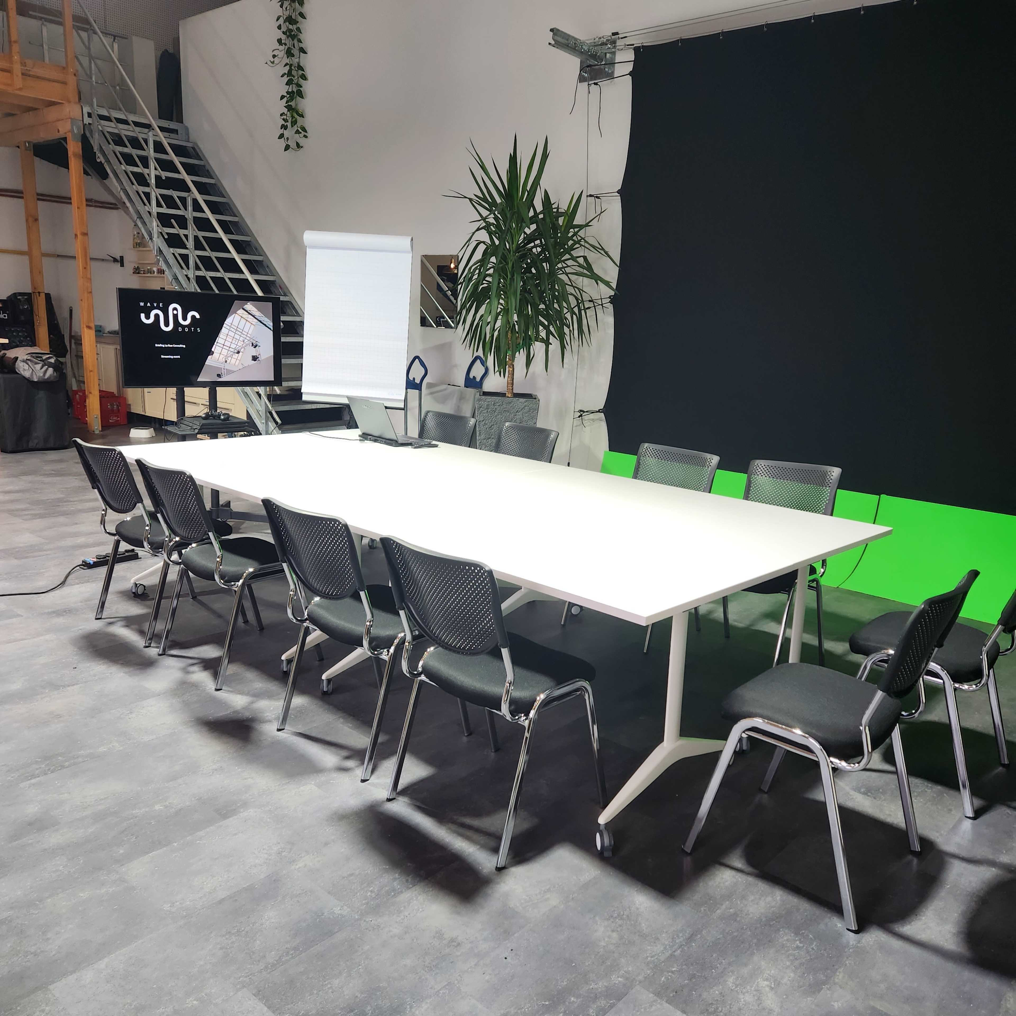 A large, white conference table surrounded by black mesh chairs is set up in a modern meeting room with a green screen backdrop.