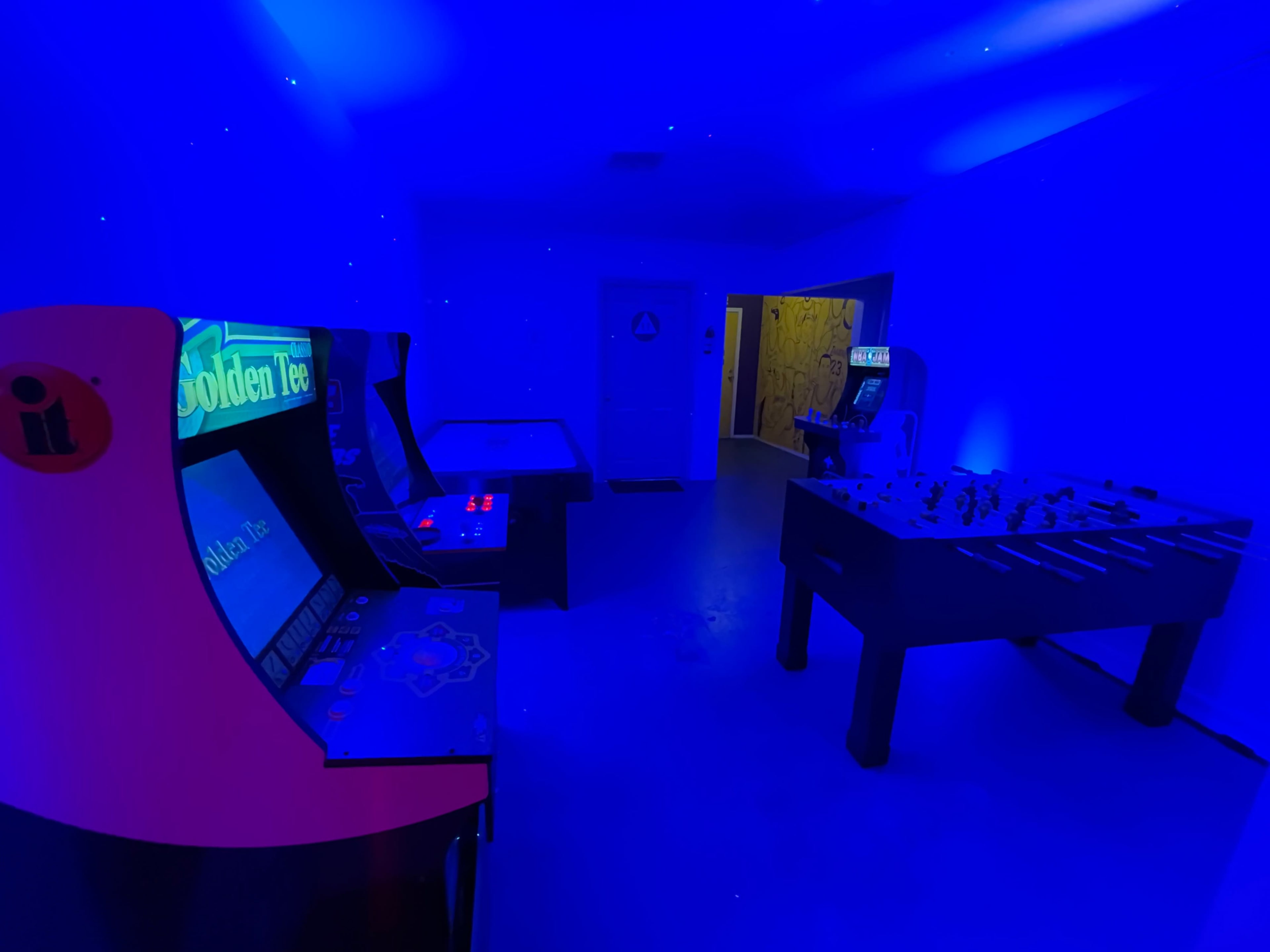 The room features retro arcade machines and a foosball table under blue lighting.