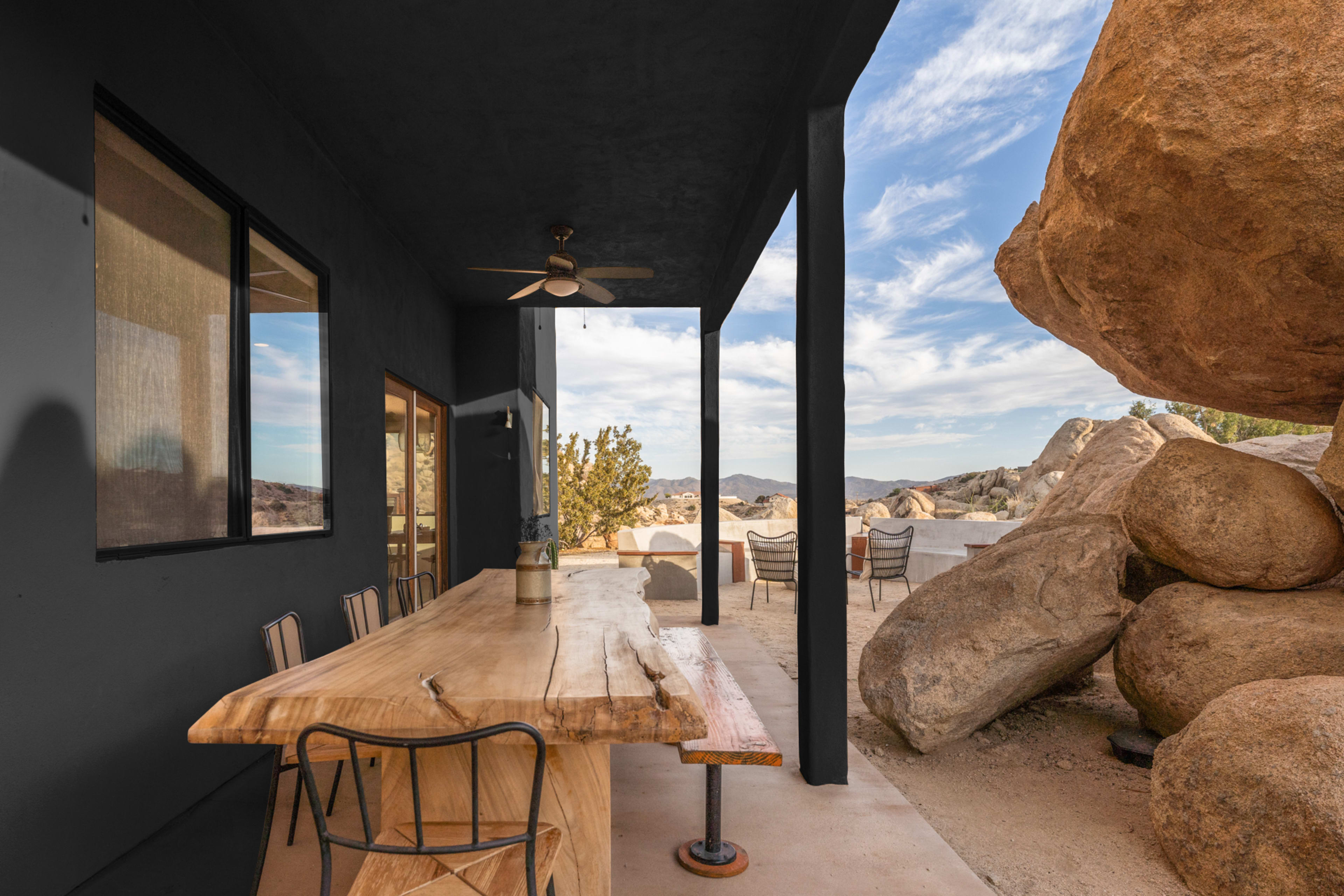 A wooden dining table is positioned on a patio beside large boulders, with a view of the surrounding landscape and sky.