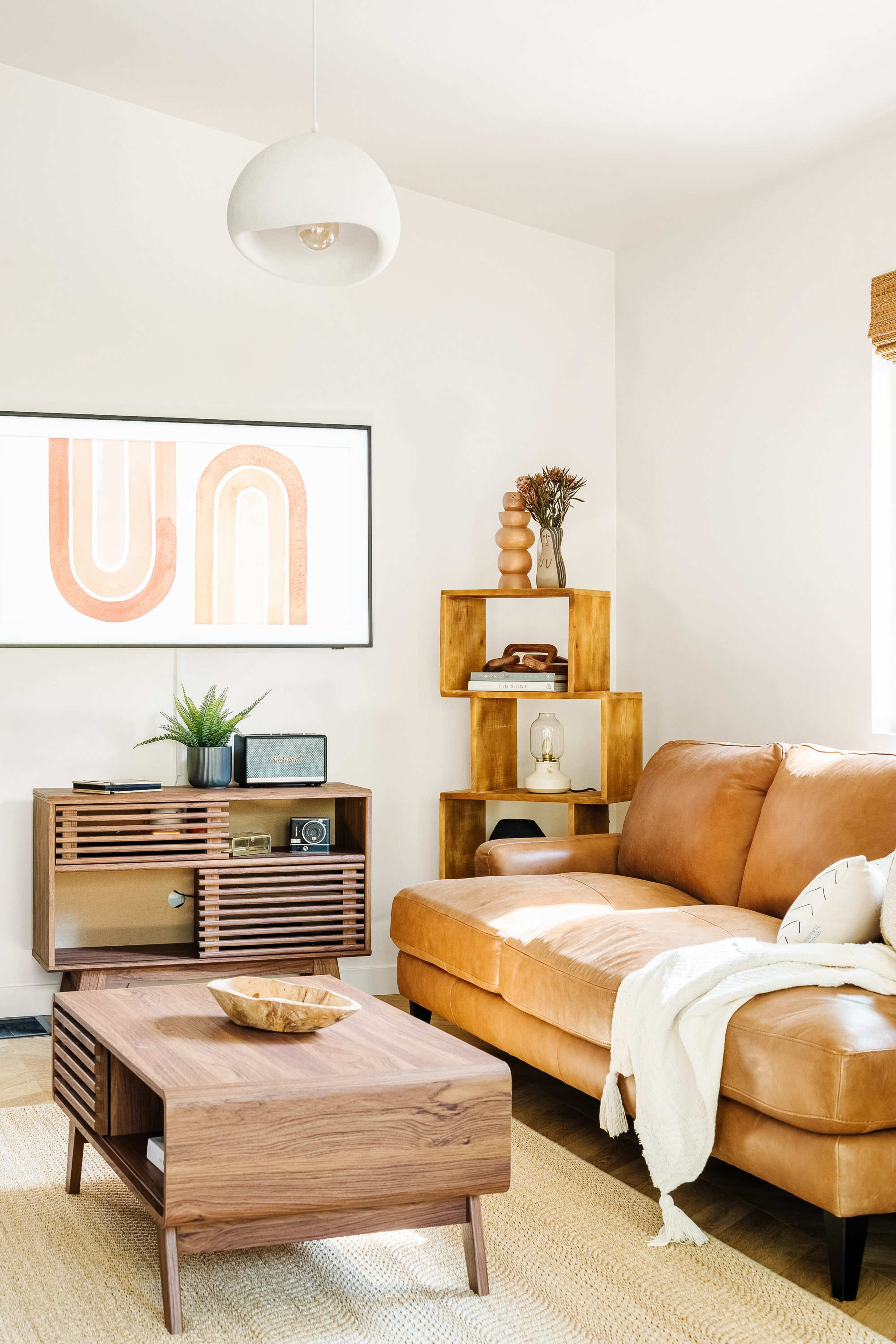 A modern living room features a brown leather sofa, a wooden coffee table, and a minimalist shelf displaying decorative items, with a framed artwork on the wall.