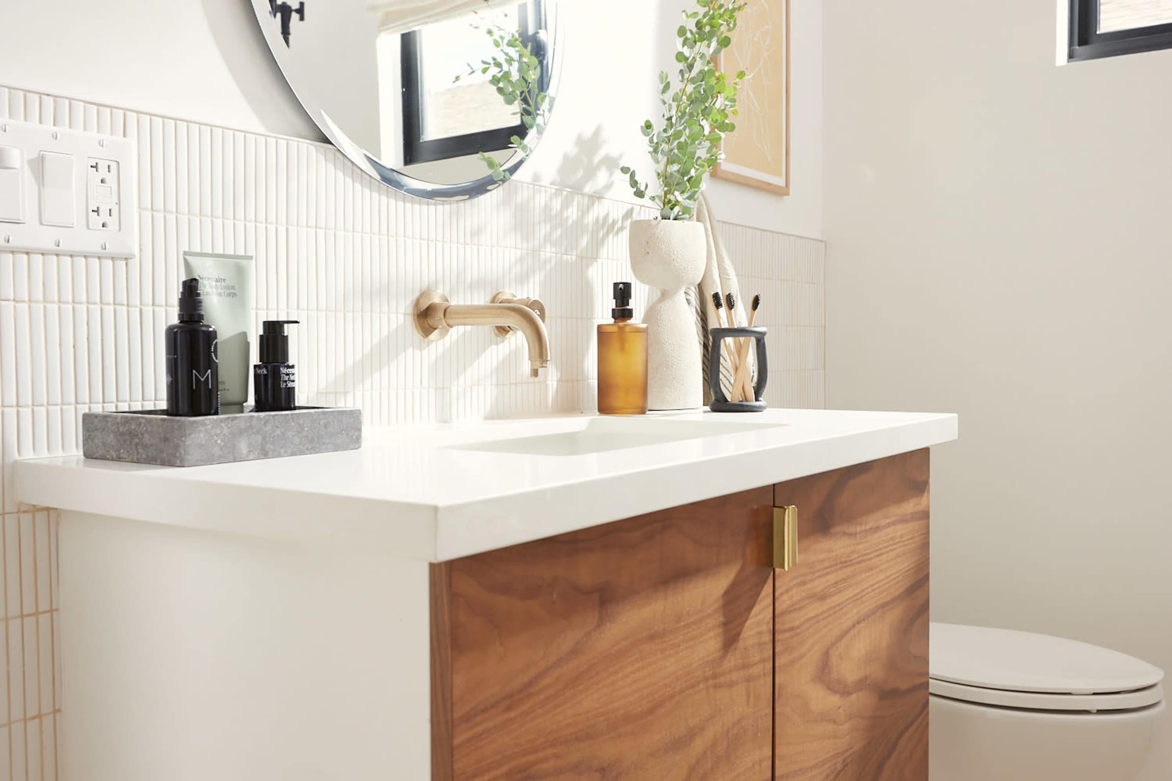 A modern bathroom features a countertop with various skincare products, a round mirror, and wooden cabinetry.
