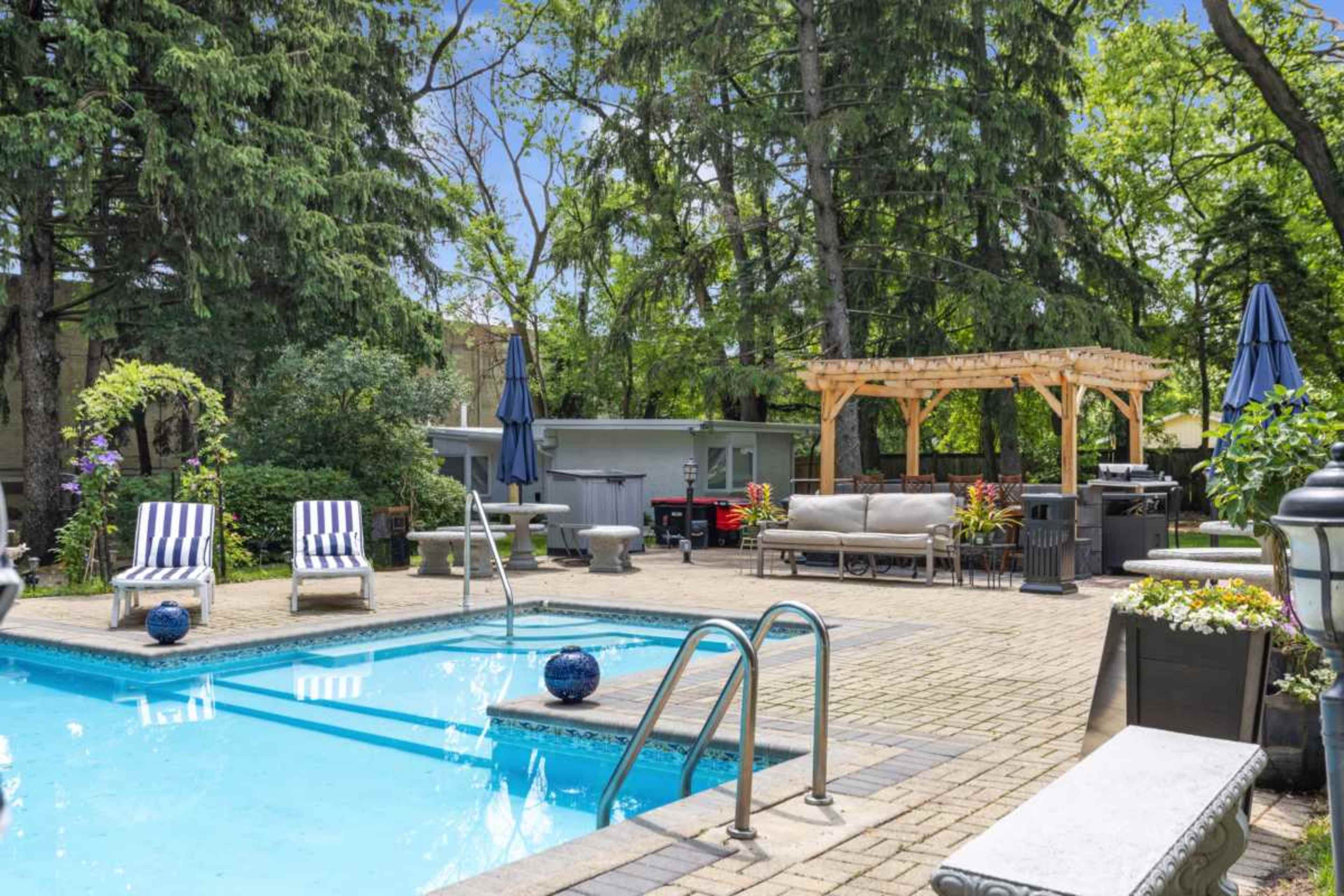 The image shows a backyard scene featuring a swimming pool, deck chairs, a shaded seating area, and surrounding greenery.