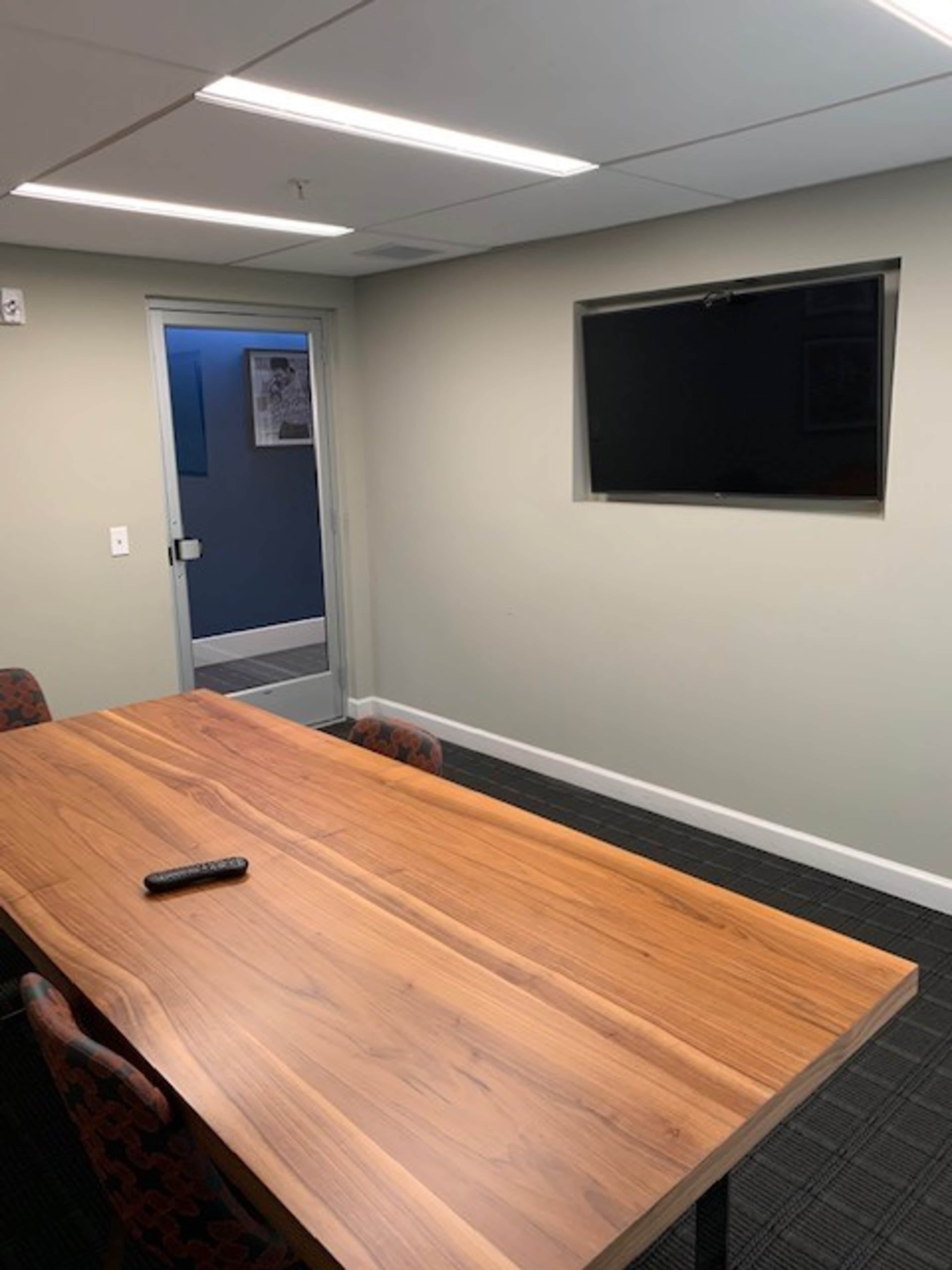 A conference room features a wooden table with a remote control on it, a mounted TV on the wall, and a glass door leading to another area.