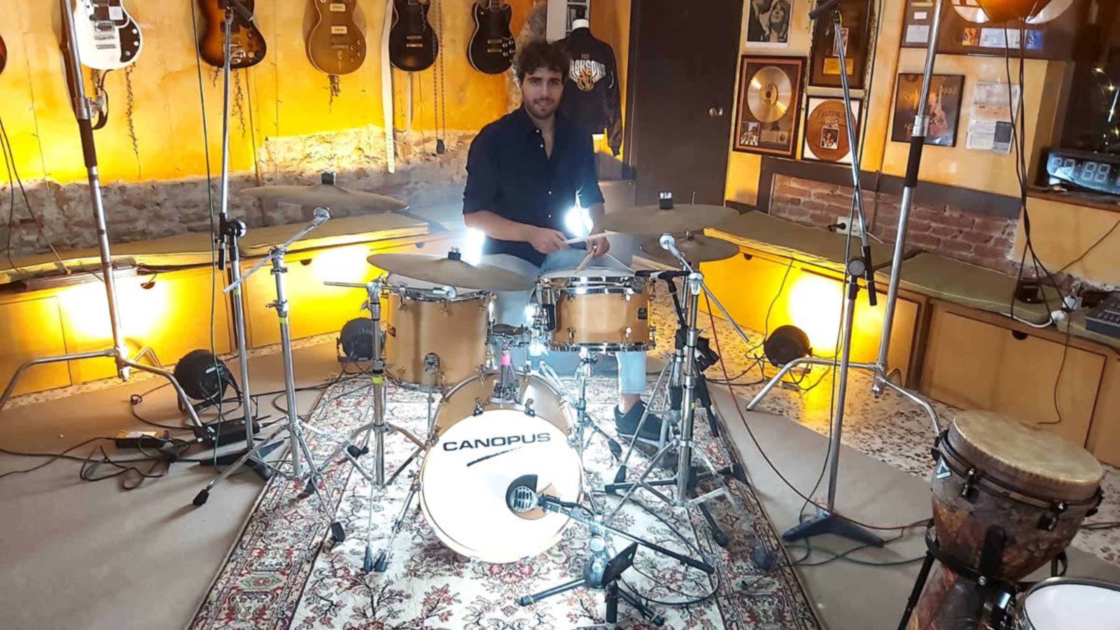 A drummer sits at a drum set surrounded by guitars and music memorabilia in a well-lit studio.