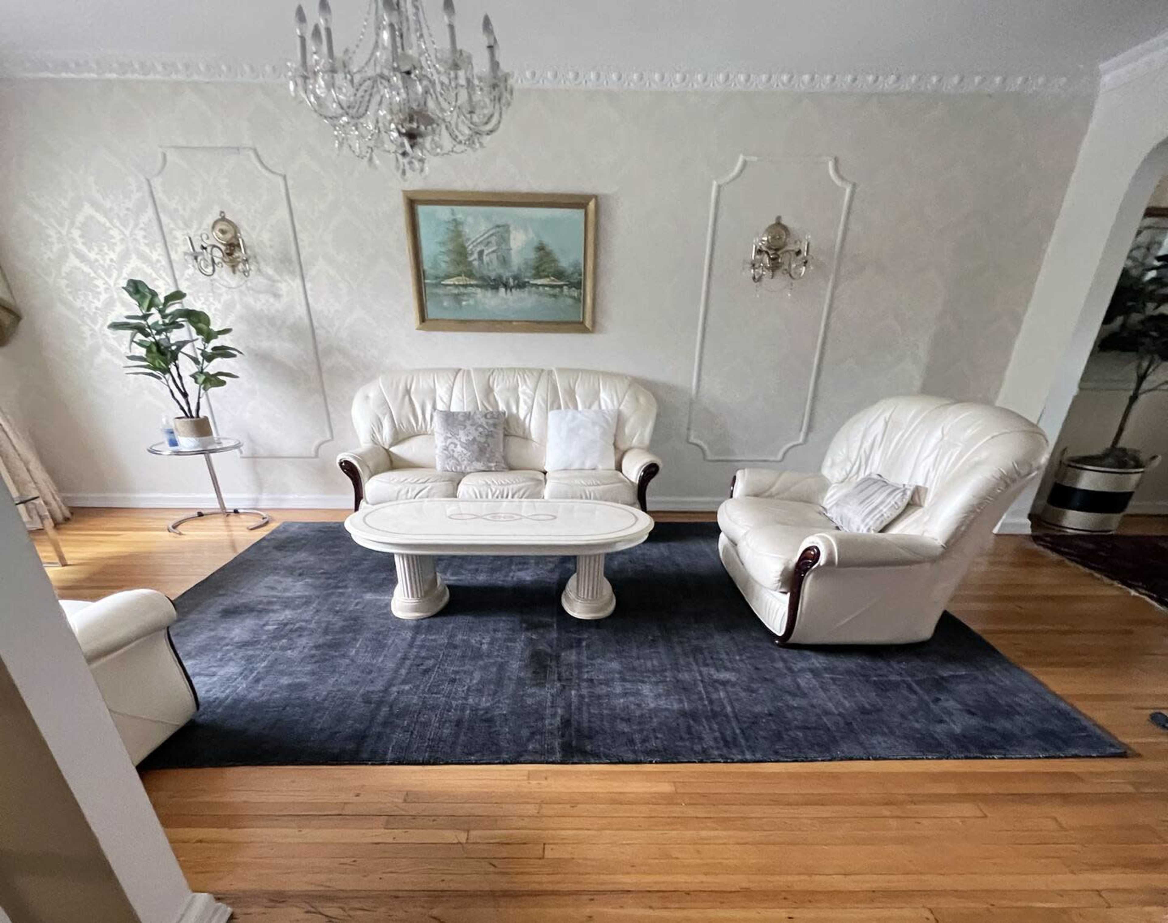 A living room with a white leather sofa and armchair, a round coffee table, and a dark area rug on wooden flooring, complemented by wall decorations and plants.
