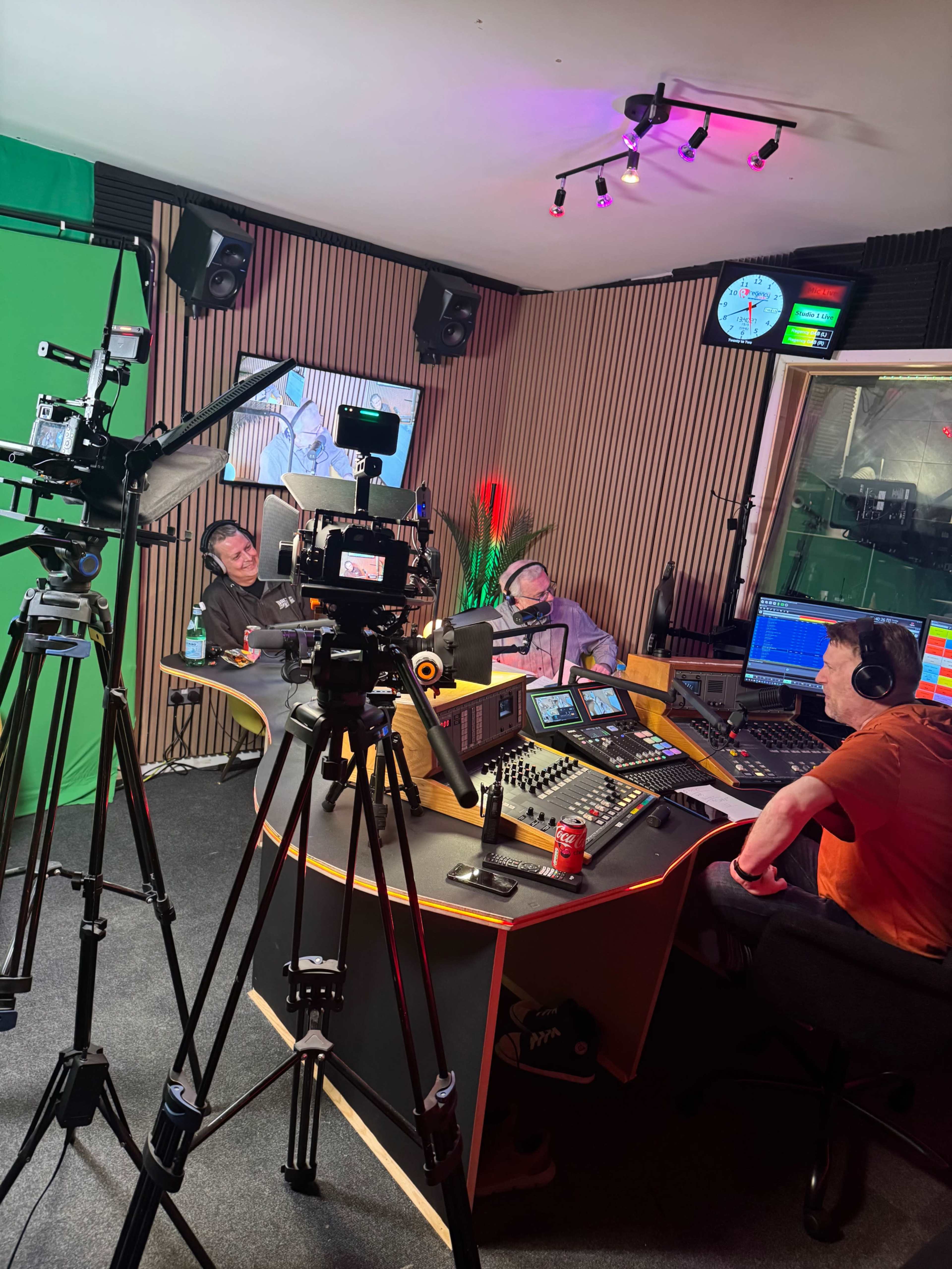 The image shows a radio studio with three people seated around a central broadcast table, surrounded by audio equipment and a green screen backdrop.