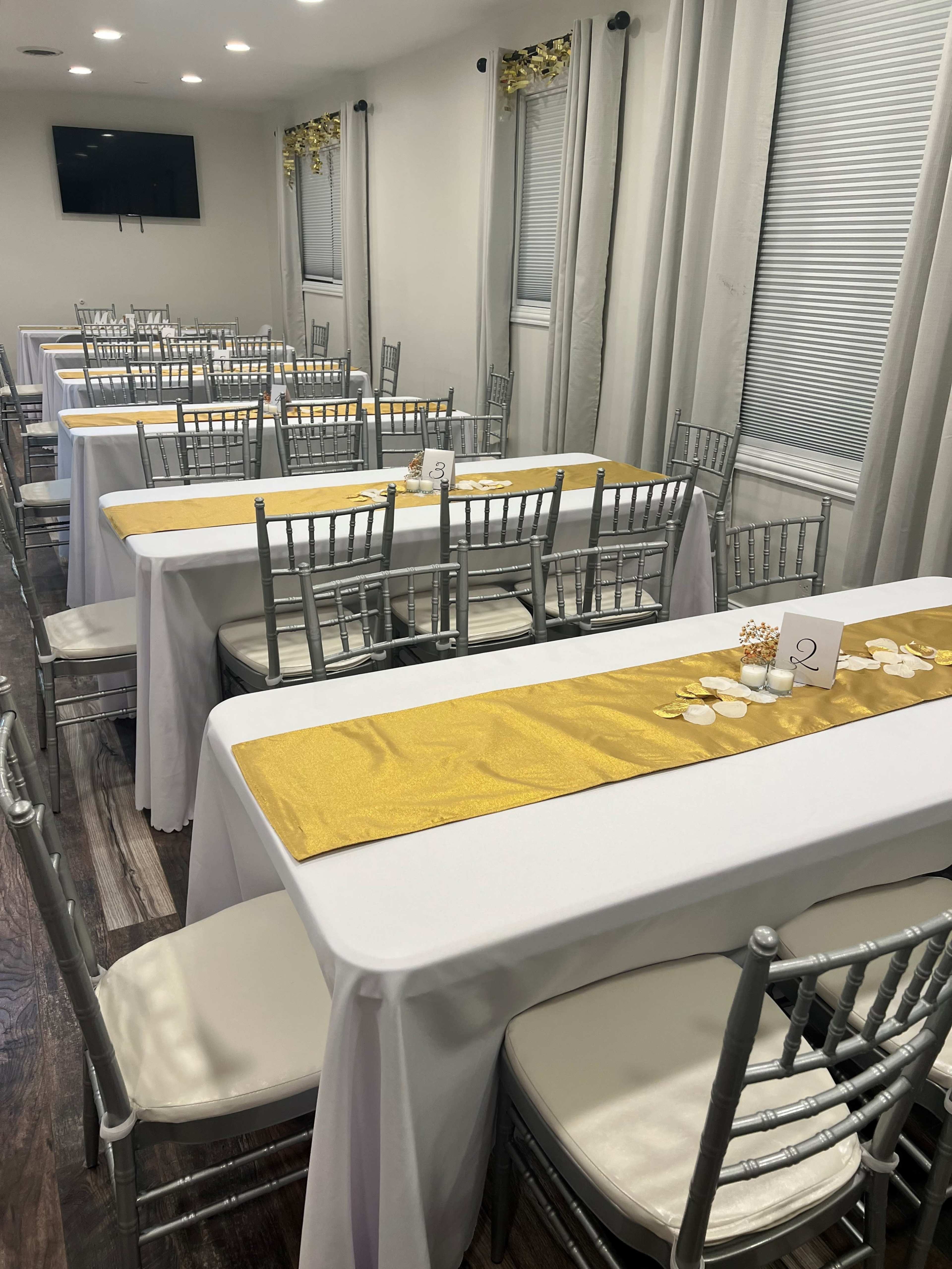 A spacious dining area set up with multiple tables covered in white tablecloths and gold runners, arranged neatly with silver chiavari chairs.