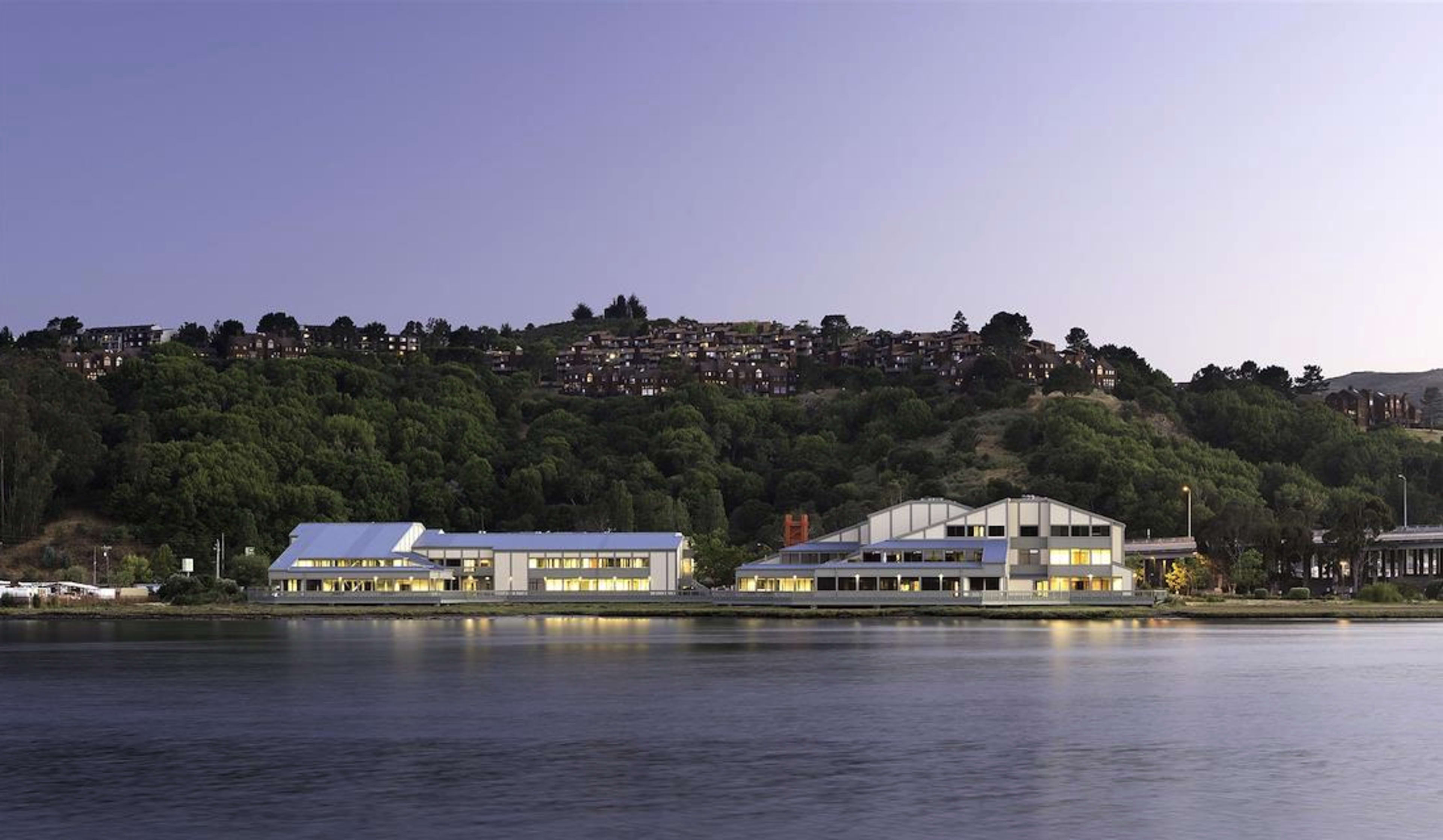 A modern building complex along a waterfront at dusk, with greenery and hillside homes in the background.