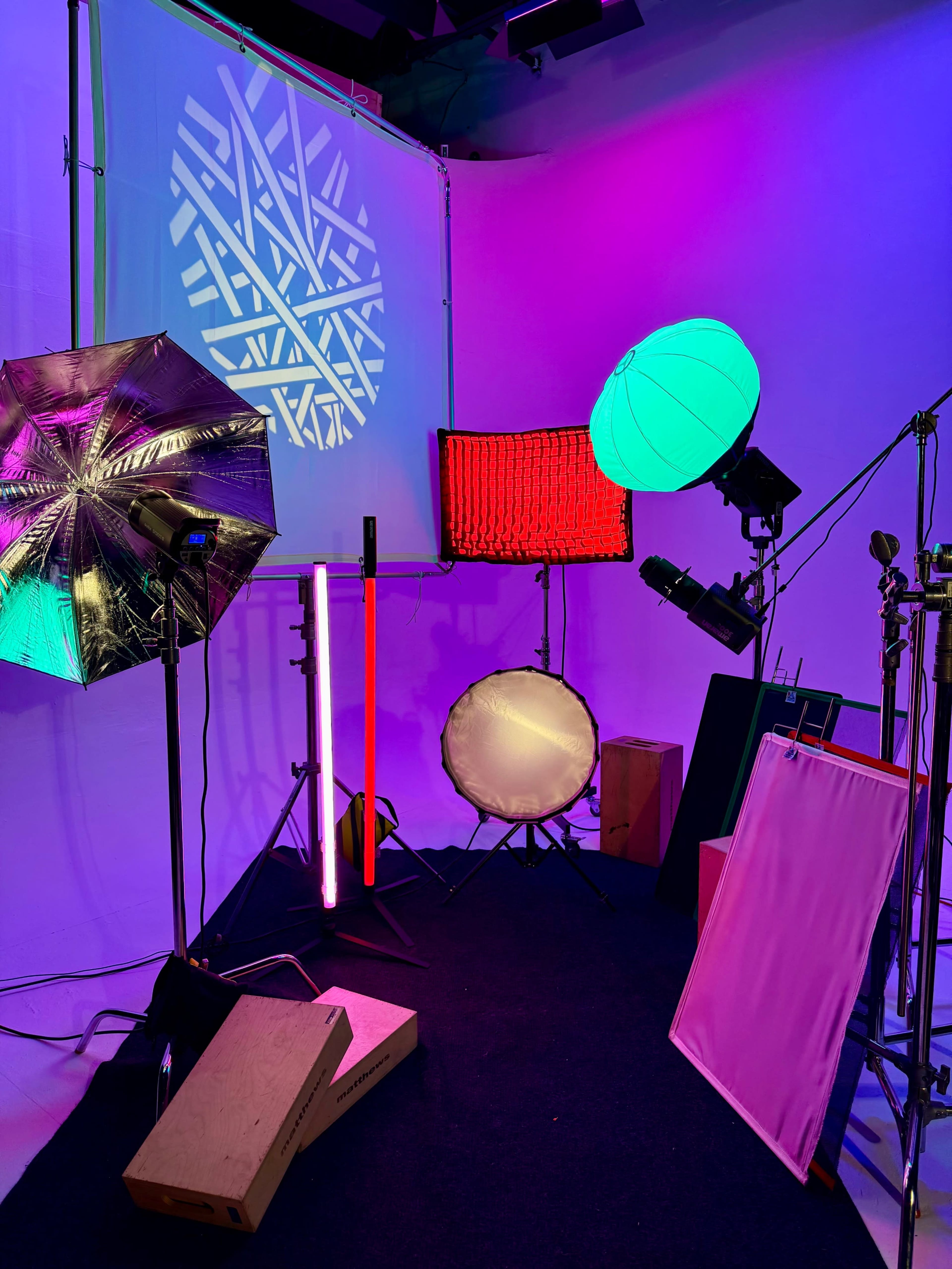 The image shows a photography studio setup with various lighting equipment, including umbrellas, softboxes, and colored lights against a white backdrop.