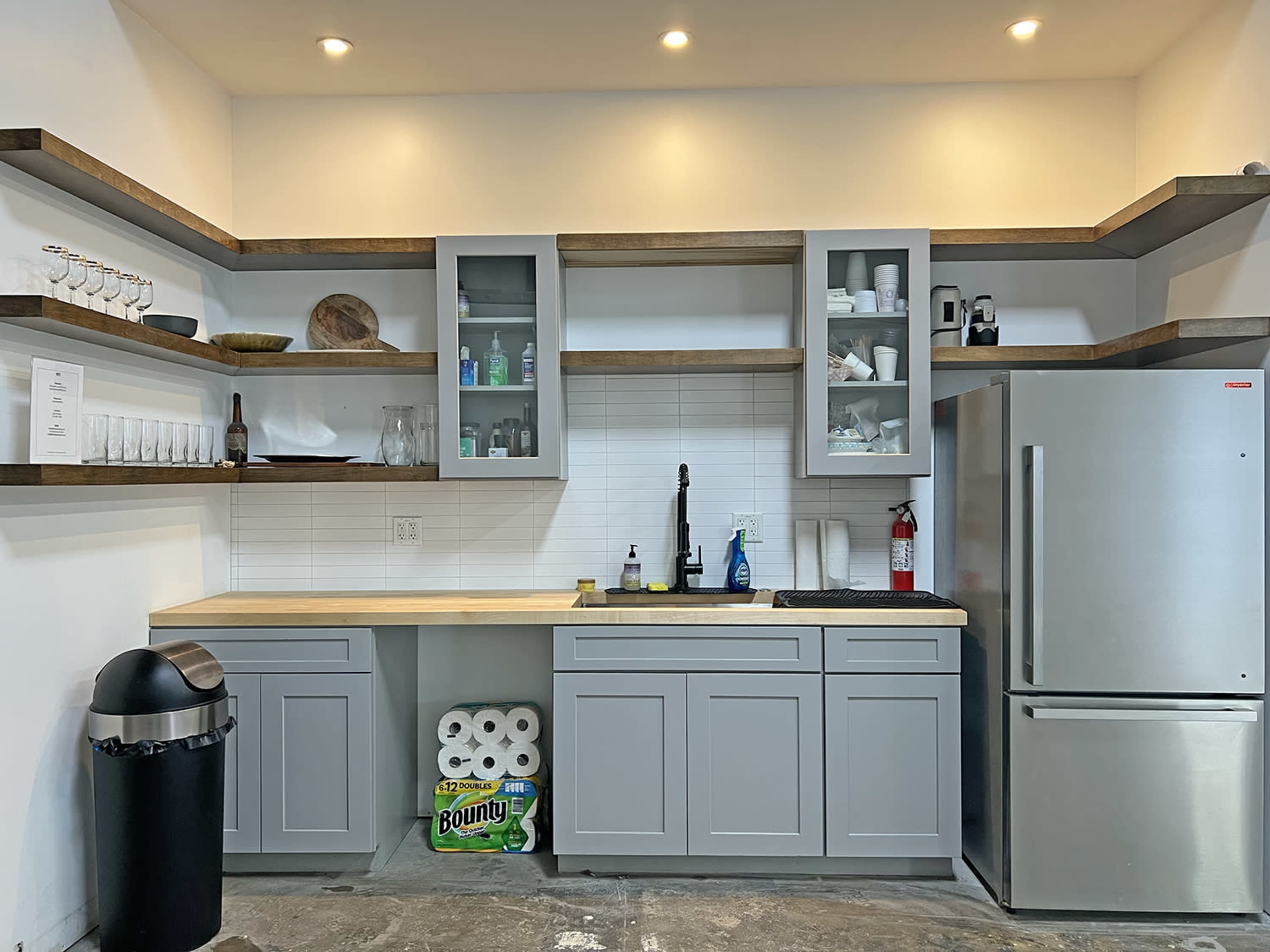 A modern kitchen with gray cabinets, a stainless steel refrigerator, and open shelving displaying various kitchen items.