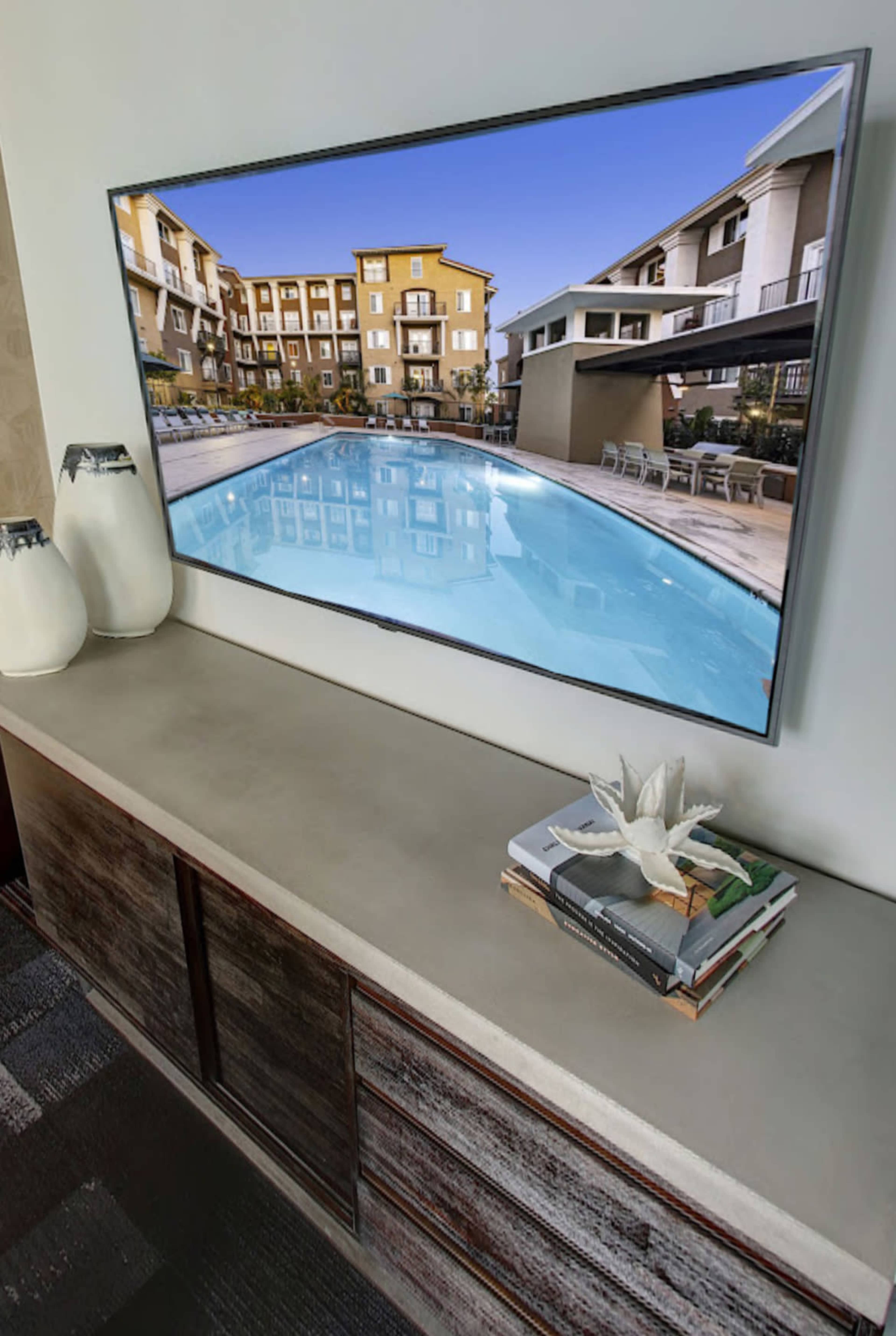 A television displaying a swimming pool scene is mounted above a concrete countertop with decorative items and magazines.