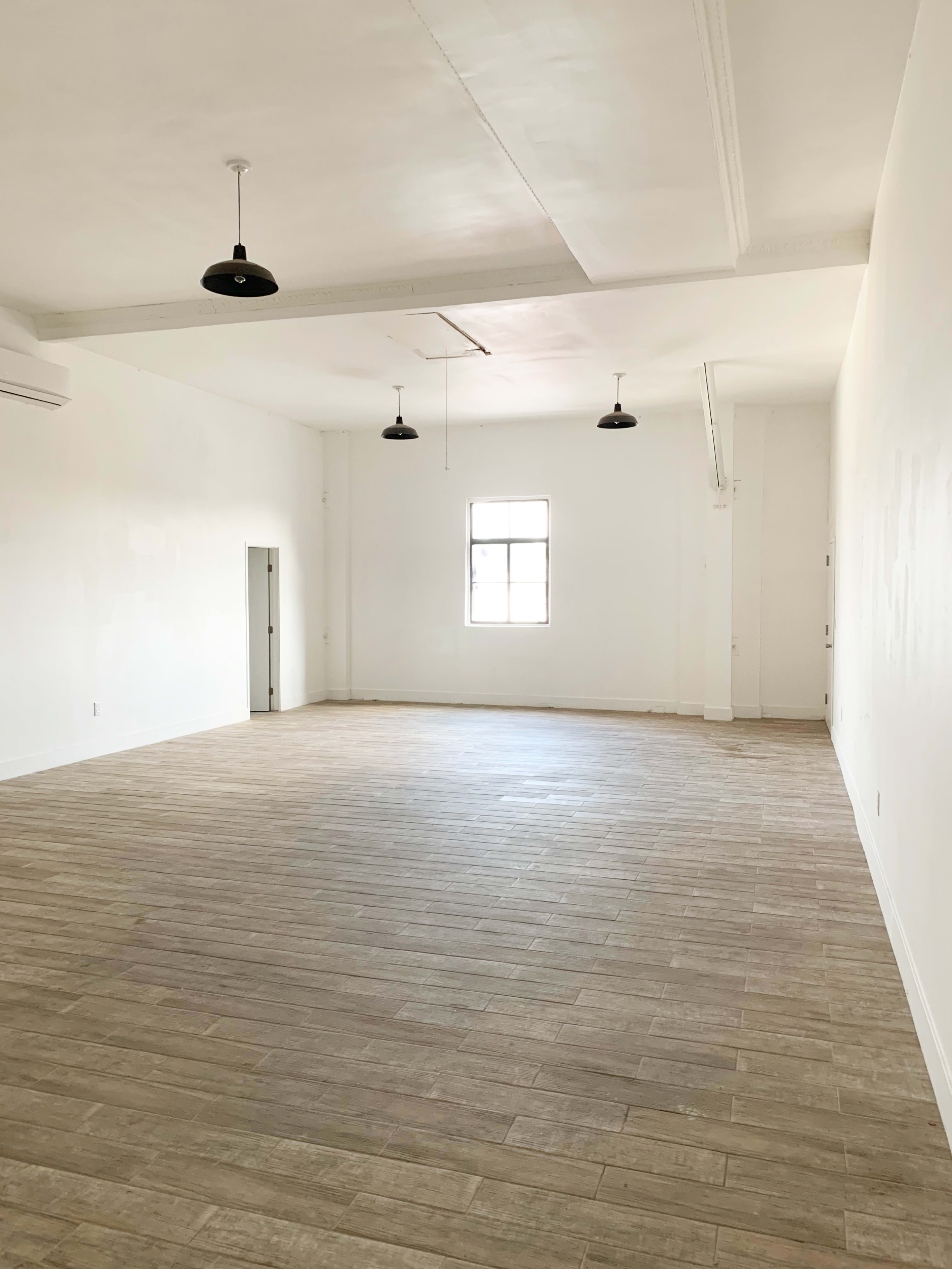 A spacious, empty room with wooden flooring and white walls, featuring a single window and pendant lights.